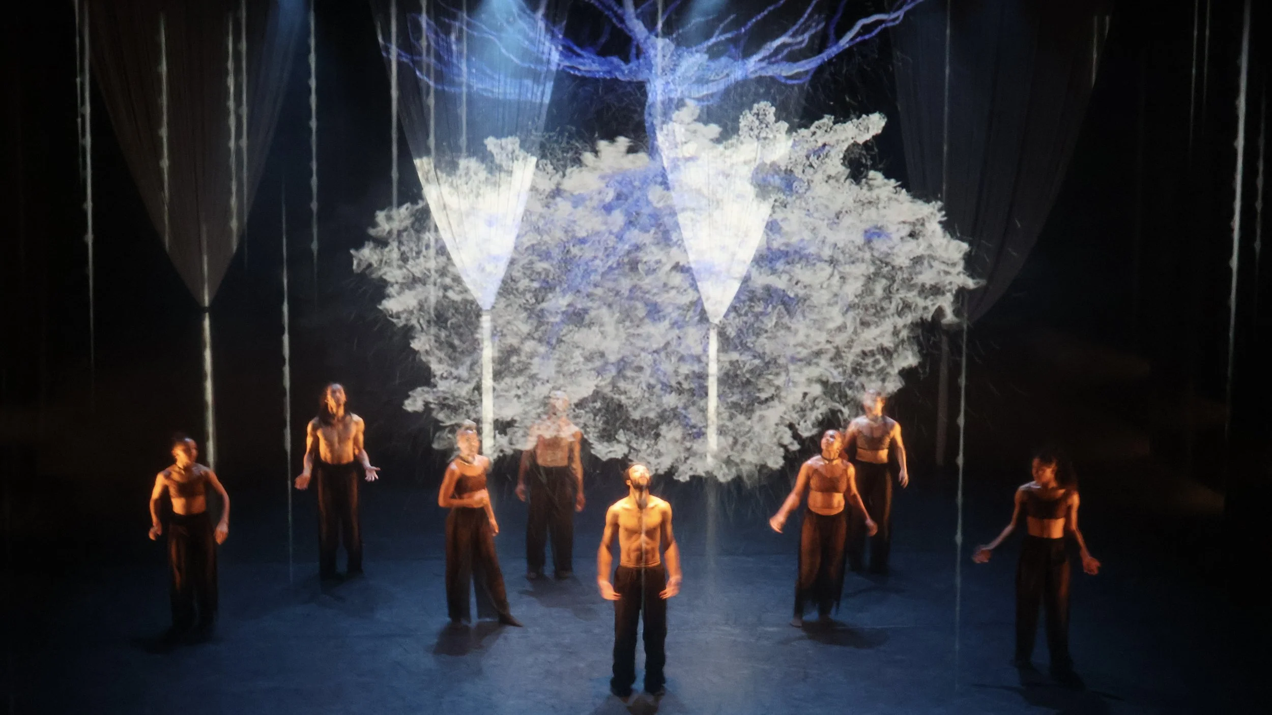 Central dancer under suspended projection tree with surrounding performers in low light in Tellus by Dickson Mbi, immersive stage design by London designer Ruby Law.