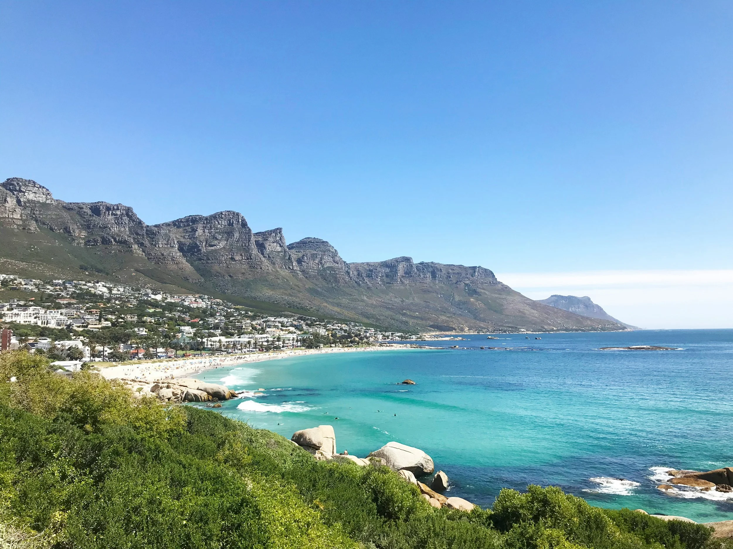 Vue d'une plage de sable blanc avec des eaux turquoise, entourée de montagnes et de verdure dense, avec un ciel clair.