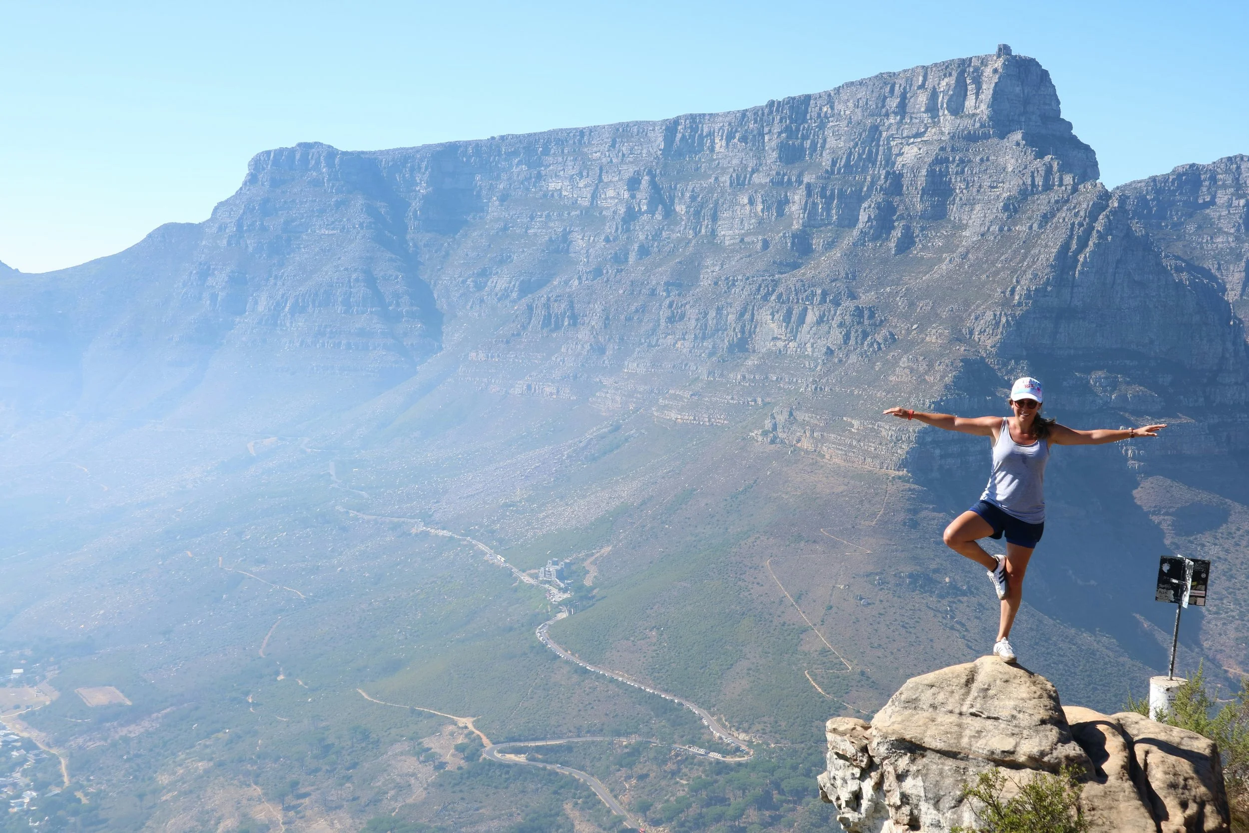 Une femme en tenue de sport, portant une casquette et des lunettes de soleil, équilibre sur une pierre au sommet d'une falaise, avec la montagne de la Table en Afrique du Sud en arrière-plan.