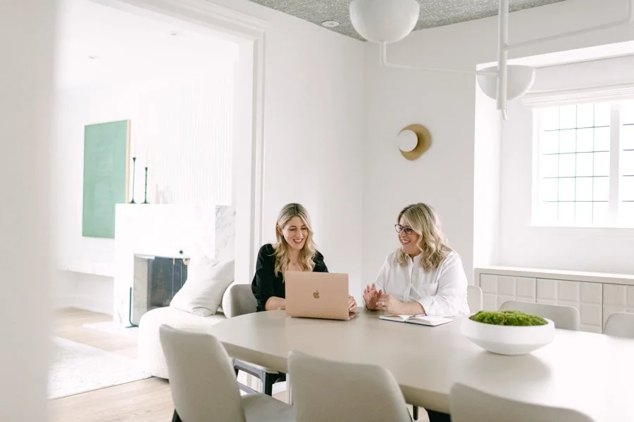 Two women sitting at a white dining table, working on a laptop and smiling, in a bright, modern, white room with large windows and minimal decor.
