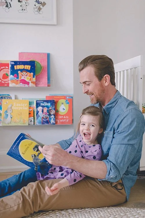 Dad in a blue shirt sitting on the floor reading to a happy child.