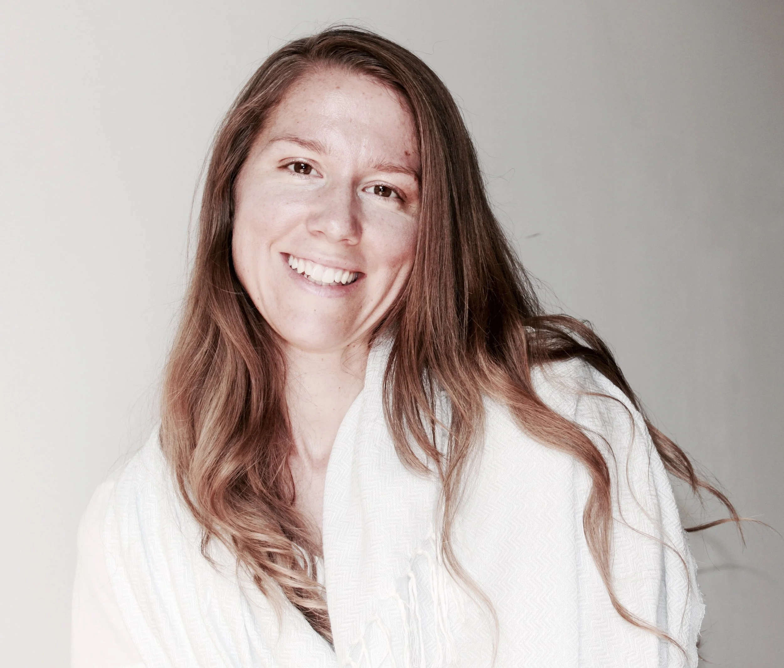 A smiling woman with long wavy reddish-brown hair wearing a white shirt with a textured pattern, posing against a plain light gray background.