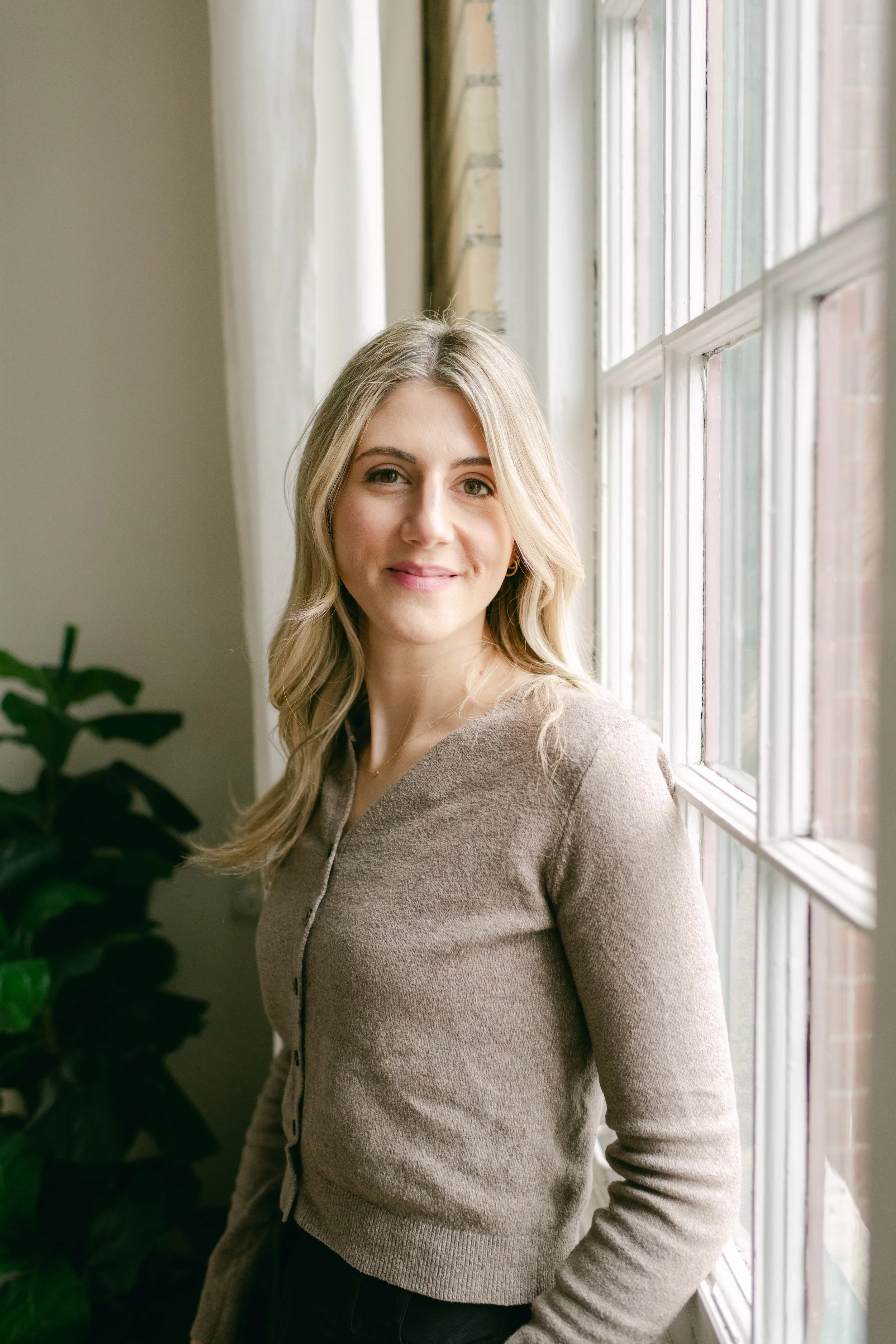 A young woman with blonde hair and a light brown sweater standing near a window with natural light.