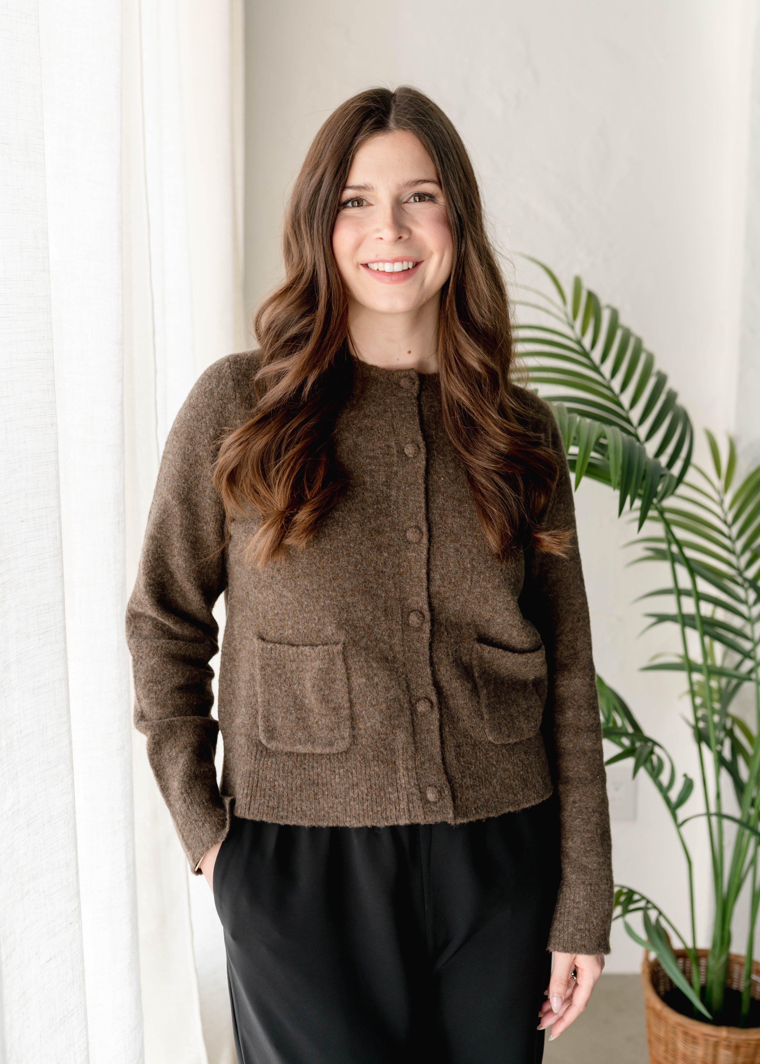 Portrait of a young woman with long brown hair, wearing a black blazer, smiling slightly, standing against a plain light-colored background.