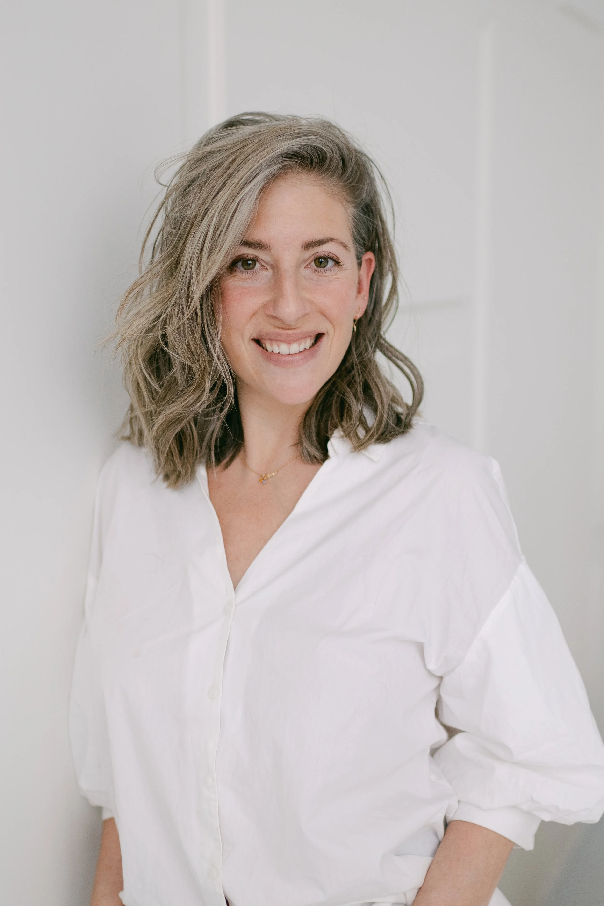 A woman with shoulder-length wavy hair wearing a white button-up shirt and small earrings, smiling at the camera
