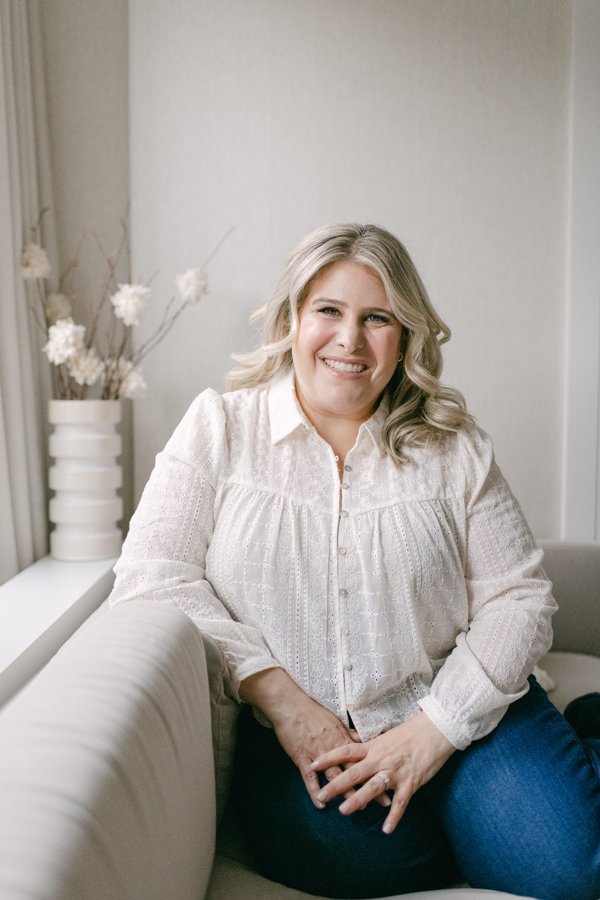A smiling woman with blonde hair wearing a white blouse sitting on a beige sofa in a bright room with a vase of white flowers on a windowsill.