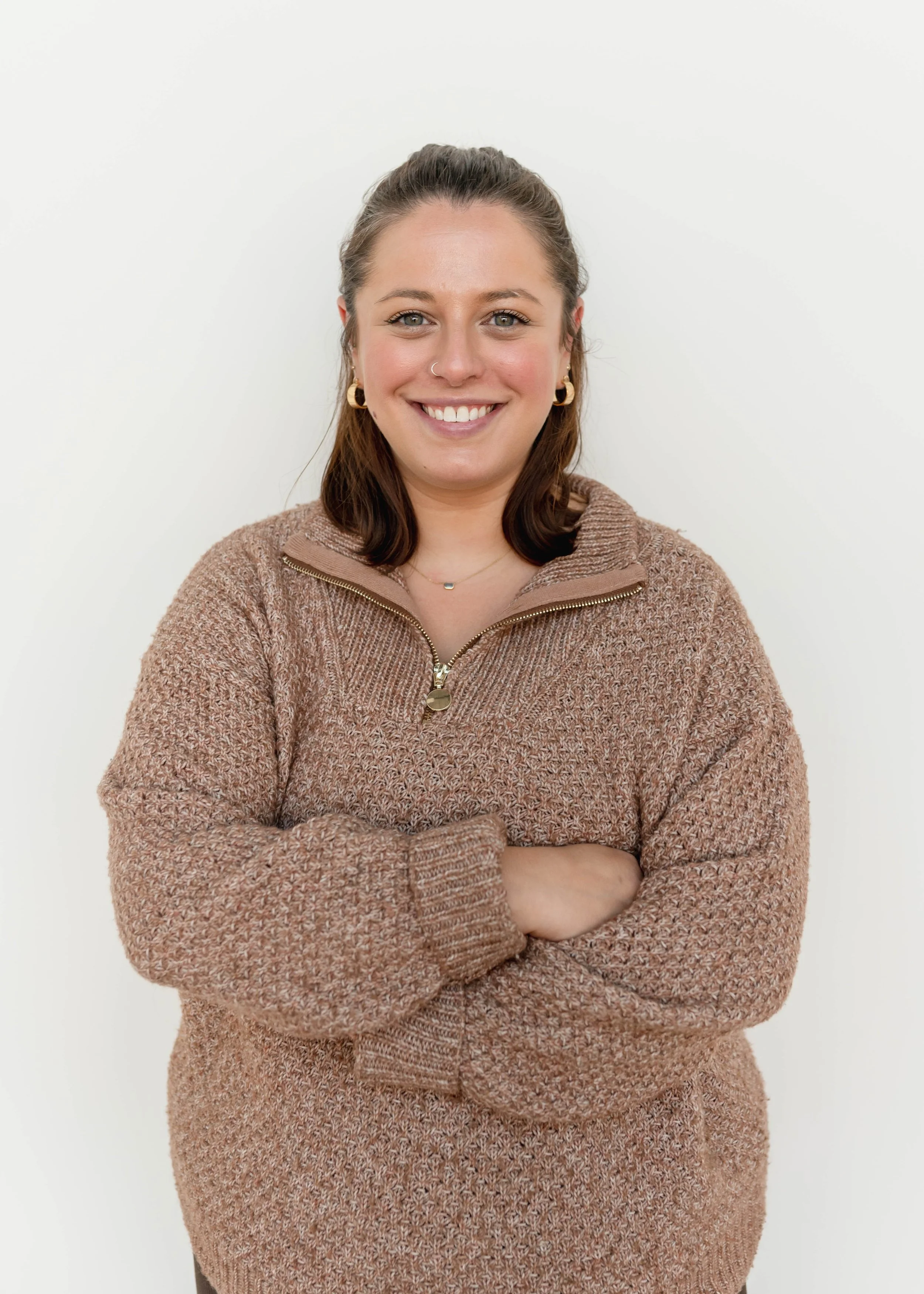 A woman with long brown hair, smiling, wearing a white t-shirt, standing in front of a white brick wall.