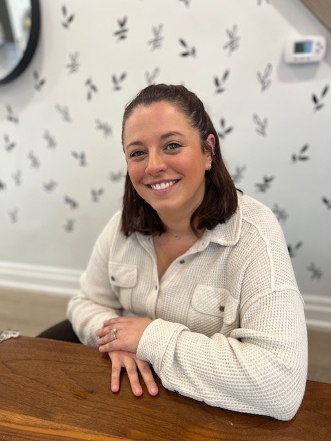A woman with dark brown hair sitting at a wooden table, smiling at the camera, with a white wall decorated with black bunny silhouette patterns behind her.