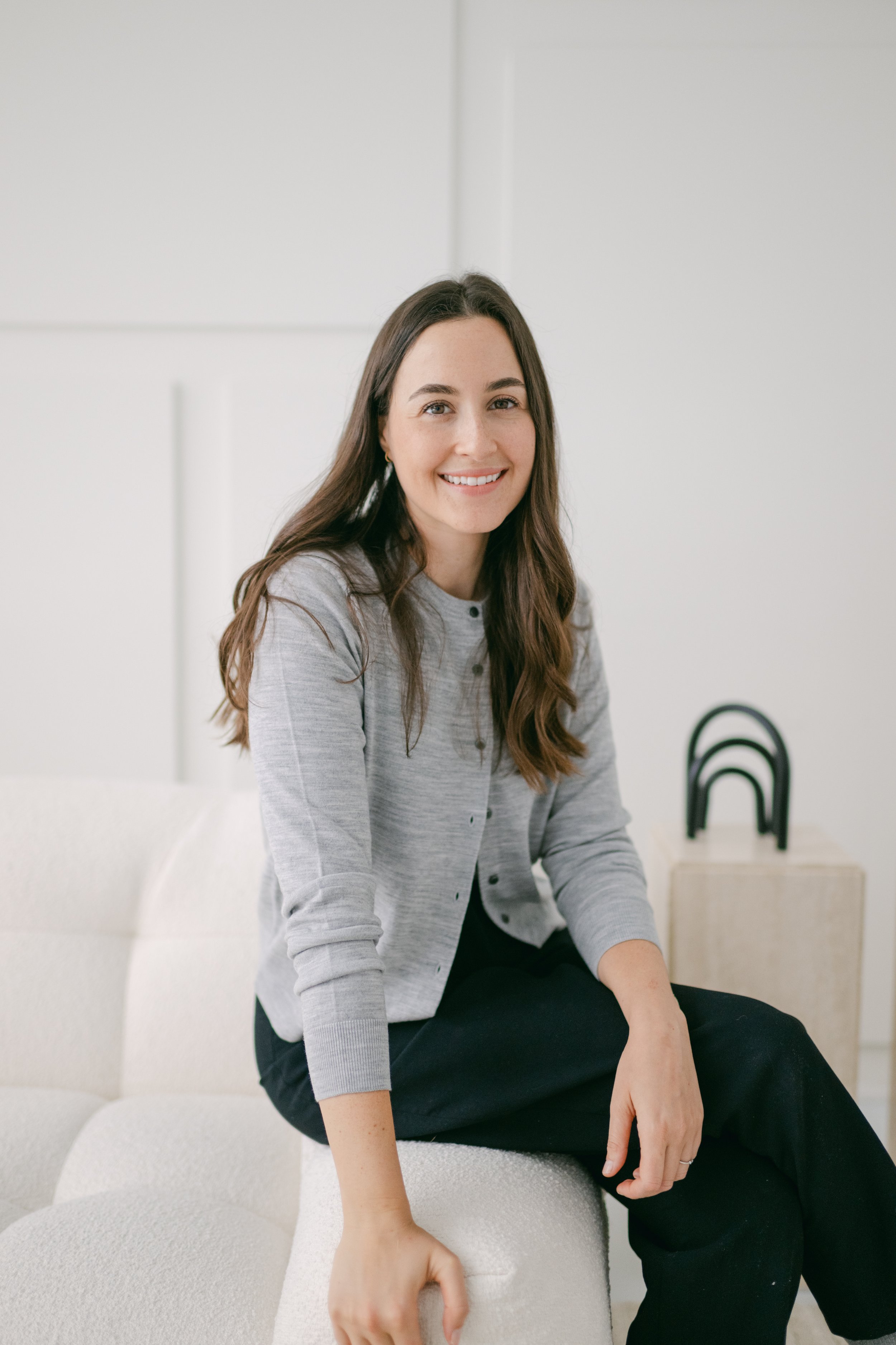 A woman with long brown hair wearing a gray cardigan and black pants sitting on a white chair in a minimalistic room.