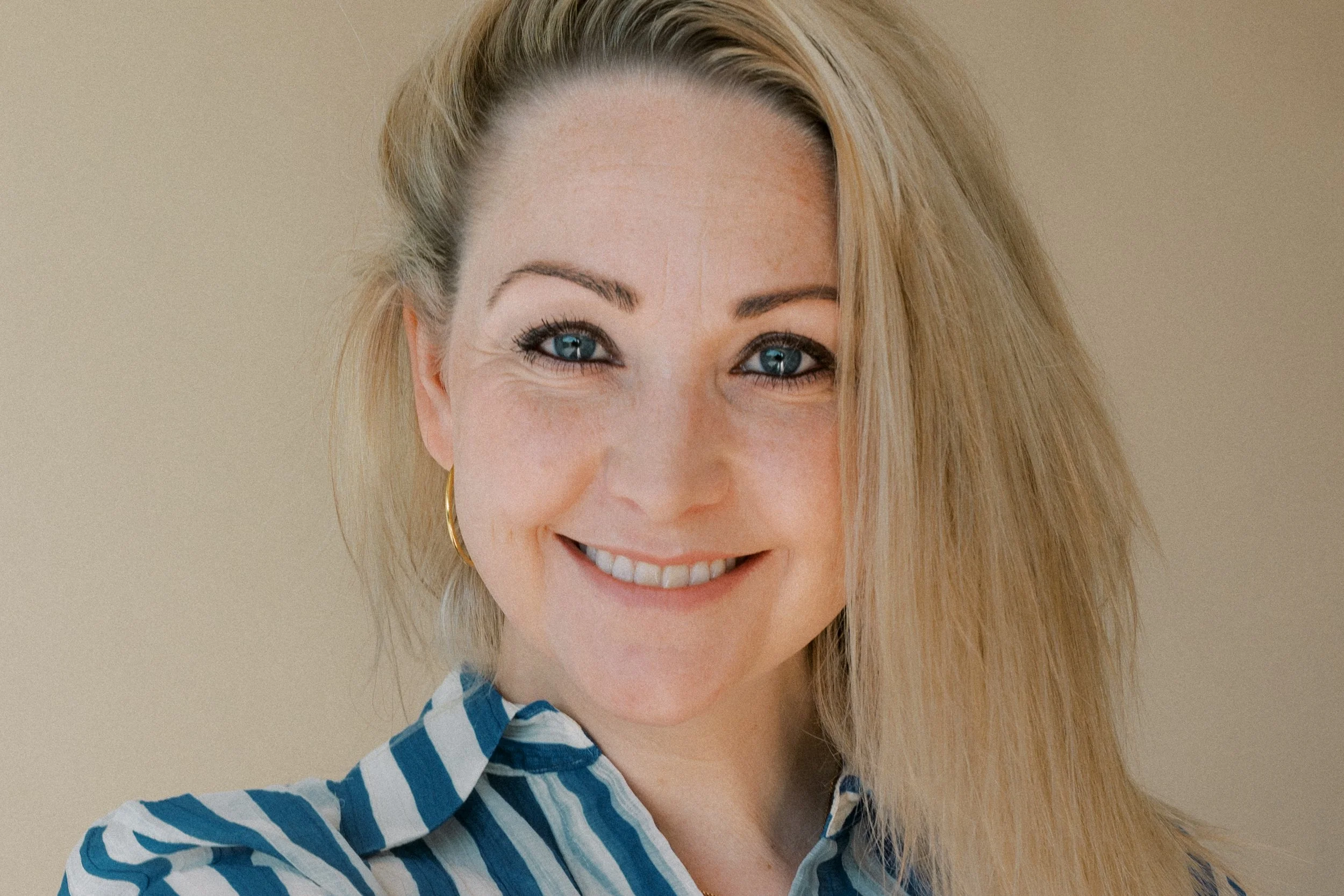 Lächelnde junge Frau mit blonden Haaren und blauen Augen, trägt einen blauen gestreiften Hemd, vor hellem Hintergrund.