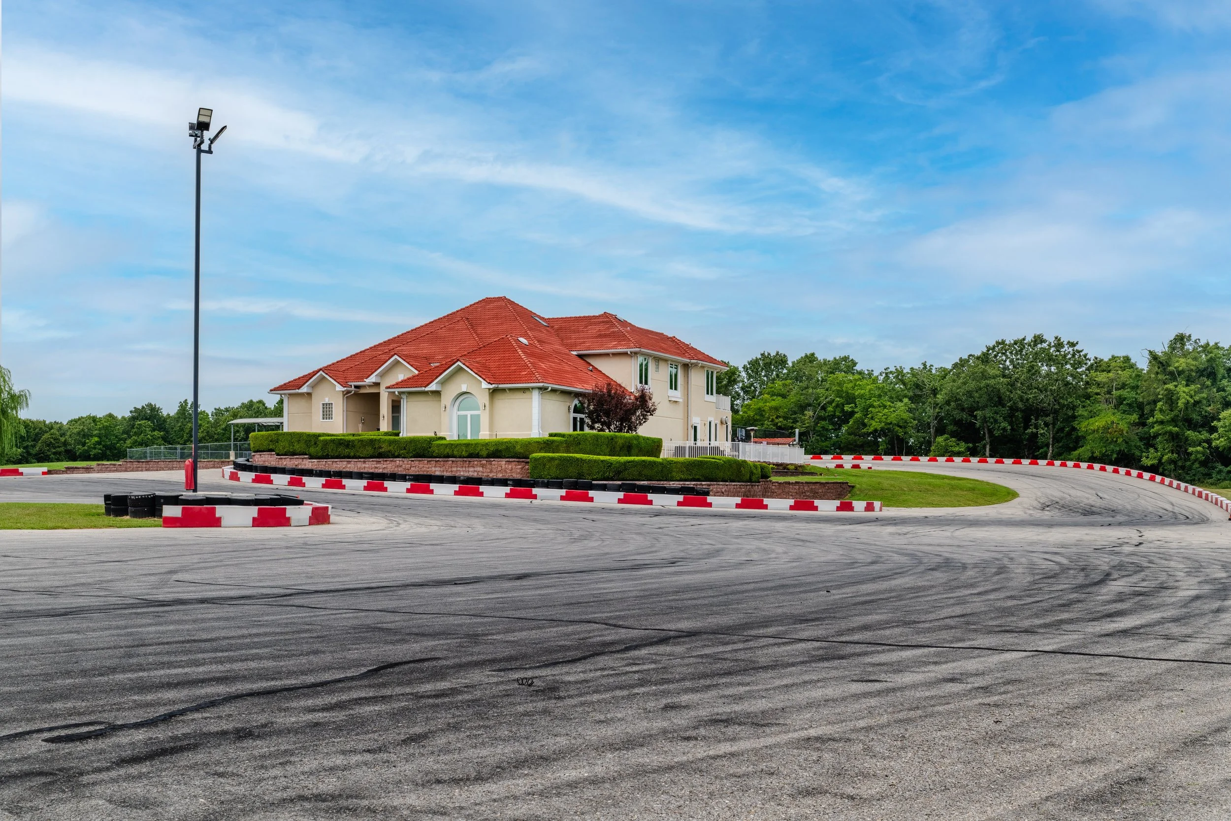 Residential building with a red-tiled roof and white walls, surrounded by trees and a well-manicured hedge, under a blue sky, near a paved area with tire marks and red-and-white barriers.