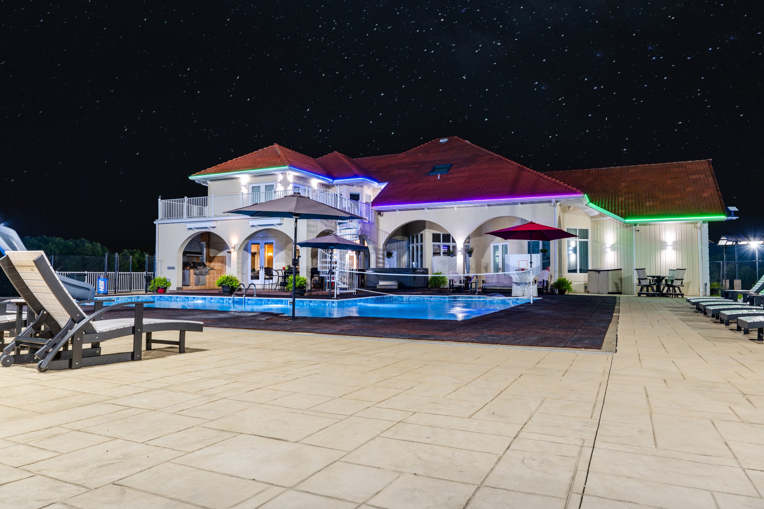 Nighttime view of a backyard with a house decorated with multicolored lights, a swimming pool, umbrellas, outdoor furniture, and a starry sky.
