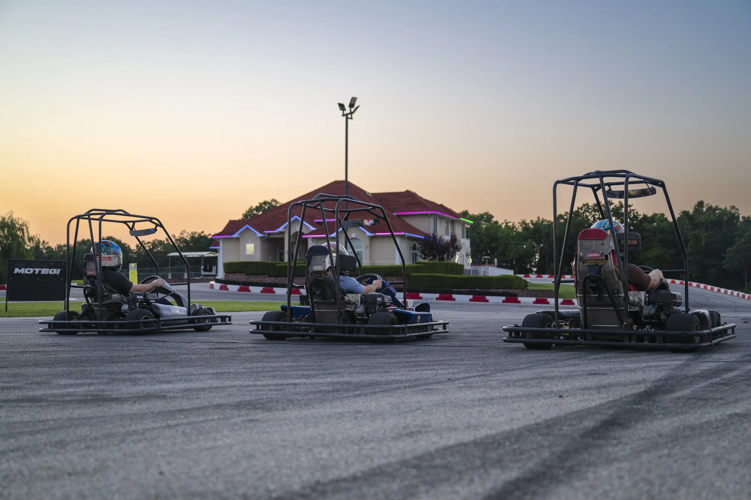 Four go-karts with riders, helmets, and safety gear, parked outside near a building with purple and green lights during sunset.