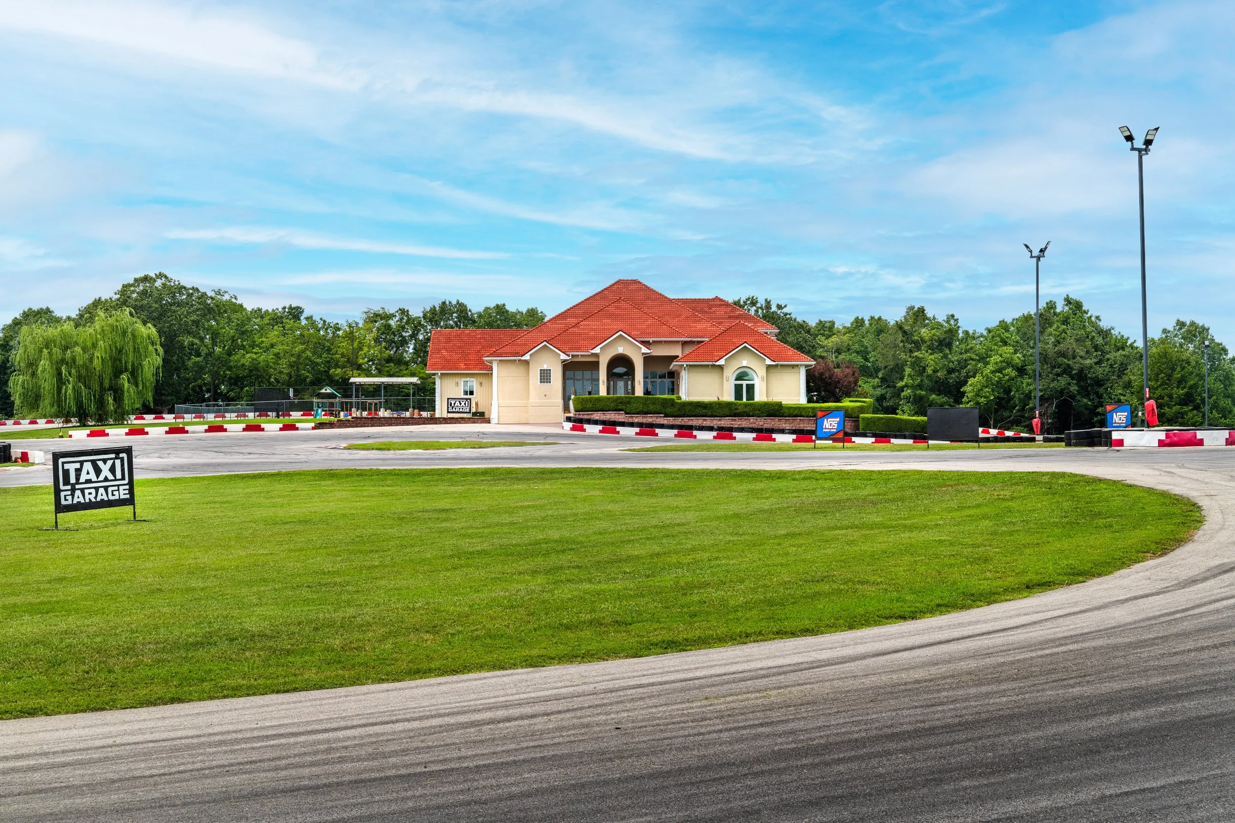 A single-story building with a red tiled roof, white walls, and an arched entrance, situated behind a lush green lawn and trees, with a clear blue sky overhead. In front, there is a paved area with signs that read 'Taxi Garage' and a couple of tall f