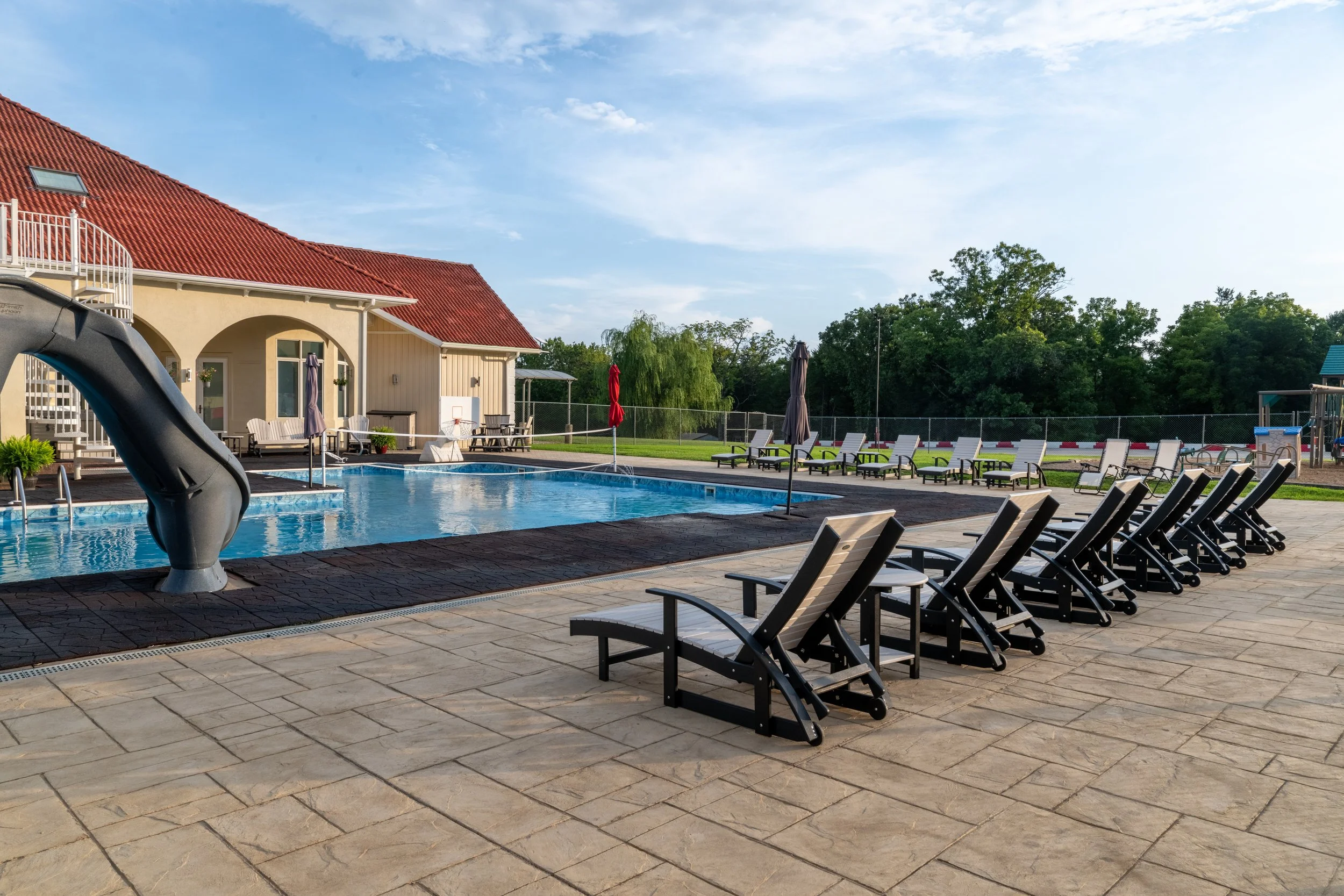 A backyard swimming pool with lounge chairs, umbrellas, and a splash slide, surrounded by a brick patio and fenced yard, with a house and trees in the background under a partly cloudy sky.