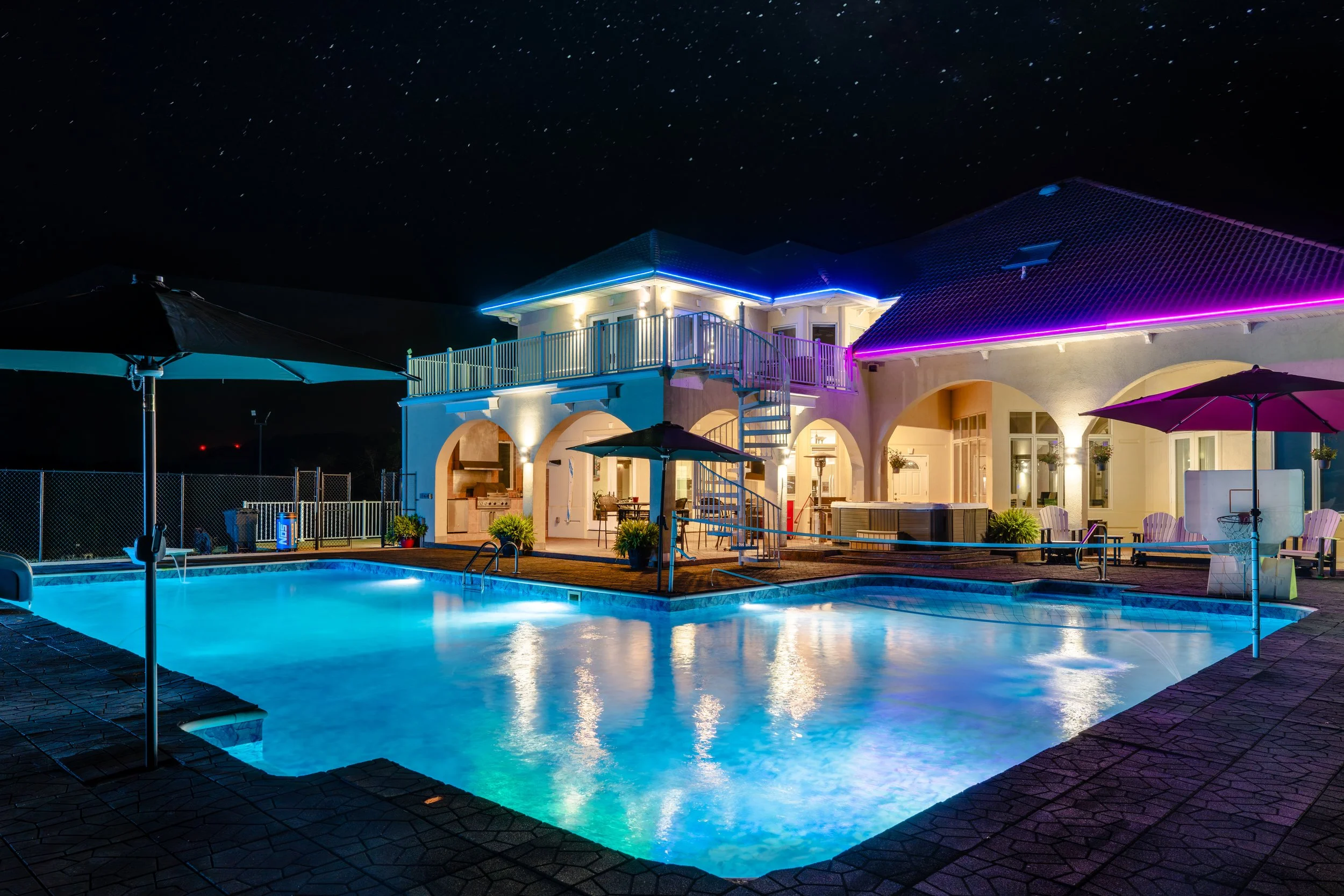 Resort house with a swimming pool under a starry night sky, illuminated with colorful neon lights along the roof edges, outdoor patio furniture, umbrellas, and potted plants.
