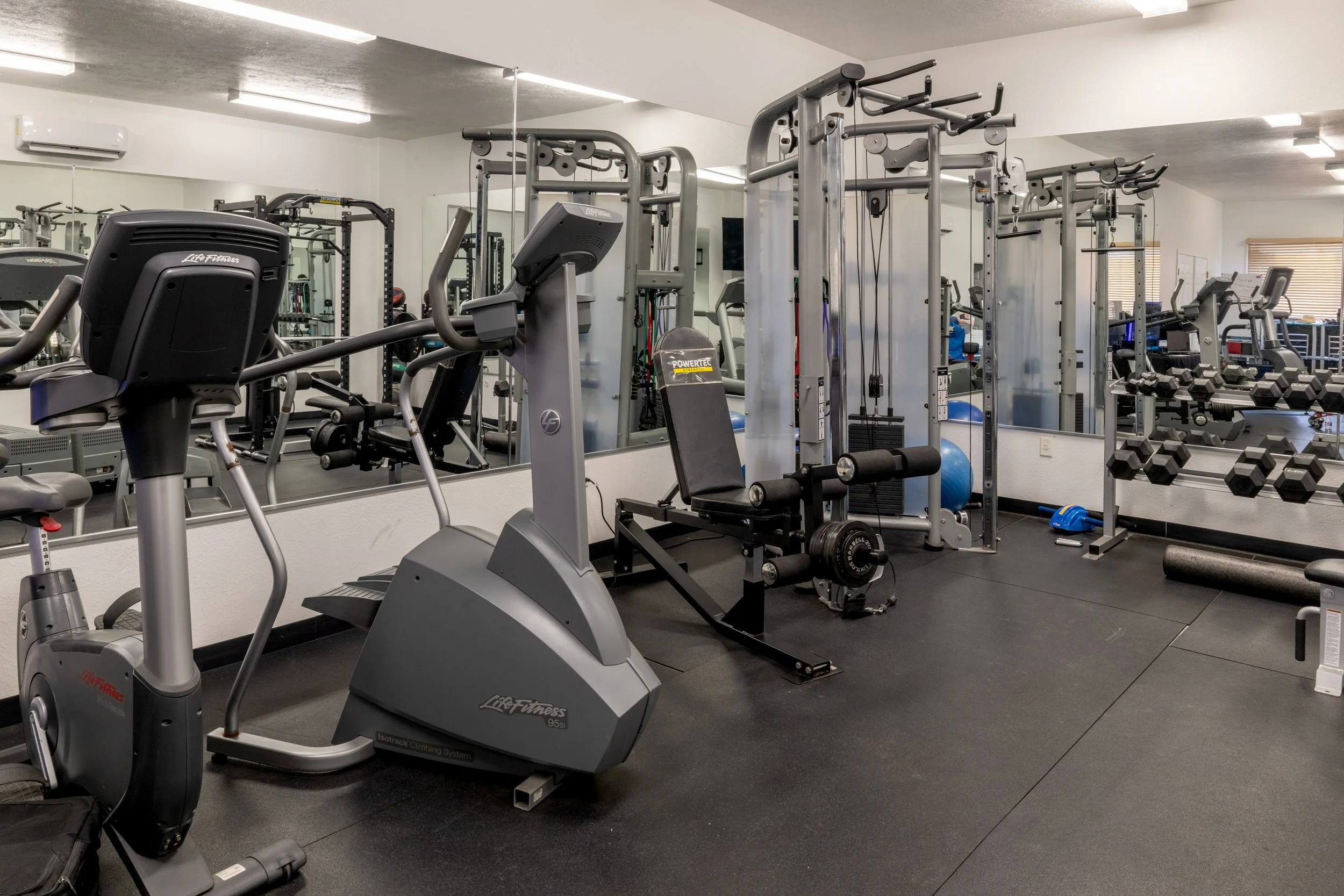 A gym with various exercise equipment including stationary bikes, weight machines, dumbbells, and stability balls in a well-lit, clean environment.