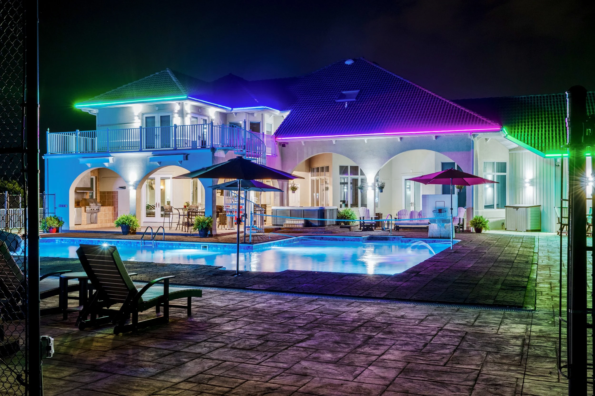 A house with a swimming pool at night, decorated with colorful neon lights outlining the roof and balcony, surrounded by patio furniture and umbrellas.