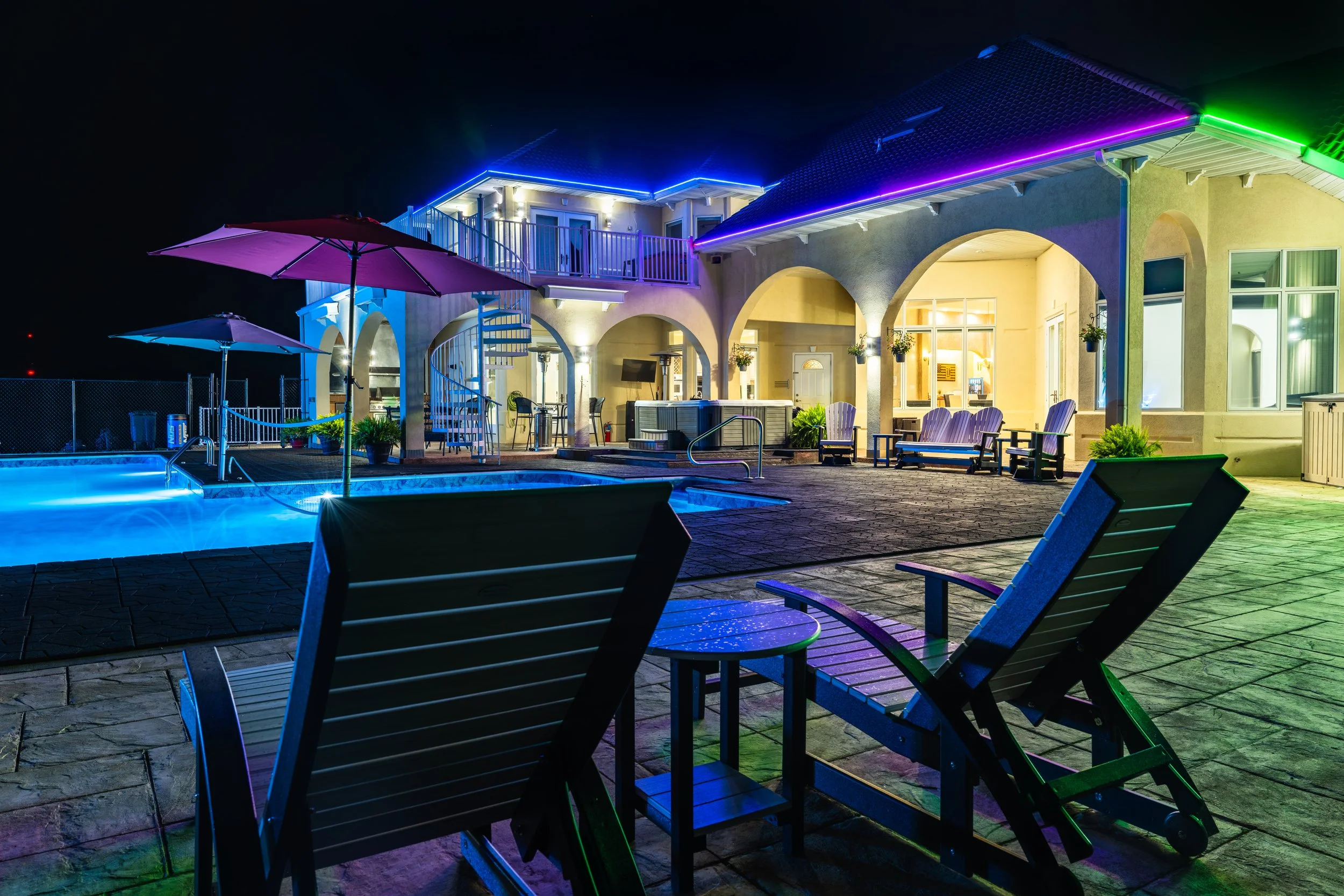 Night view of a house with colorful exterior LED lights, a swimming pool with lounge chairs, umbrellas, and an outdoor entertainment area.