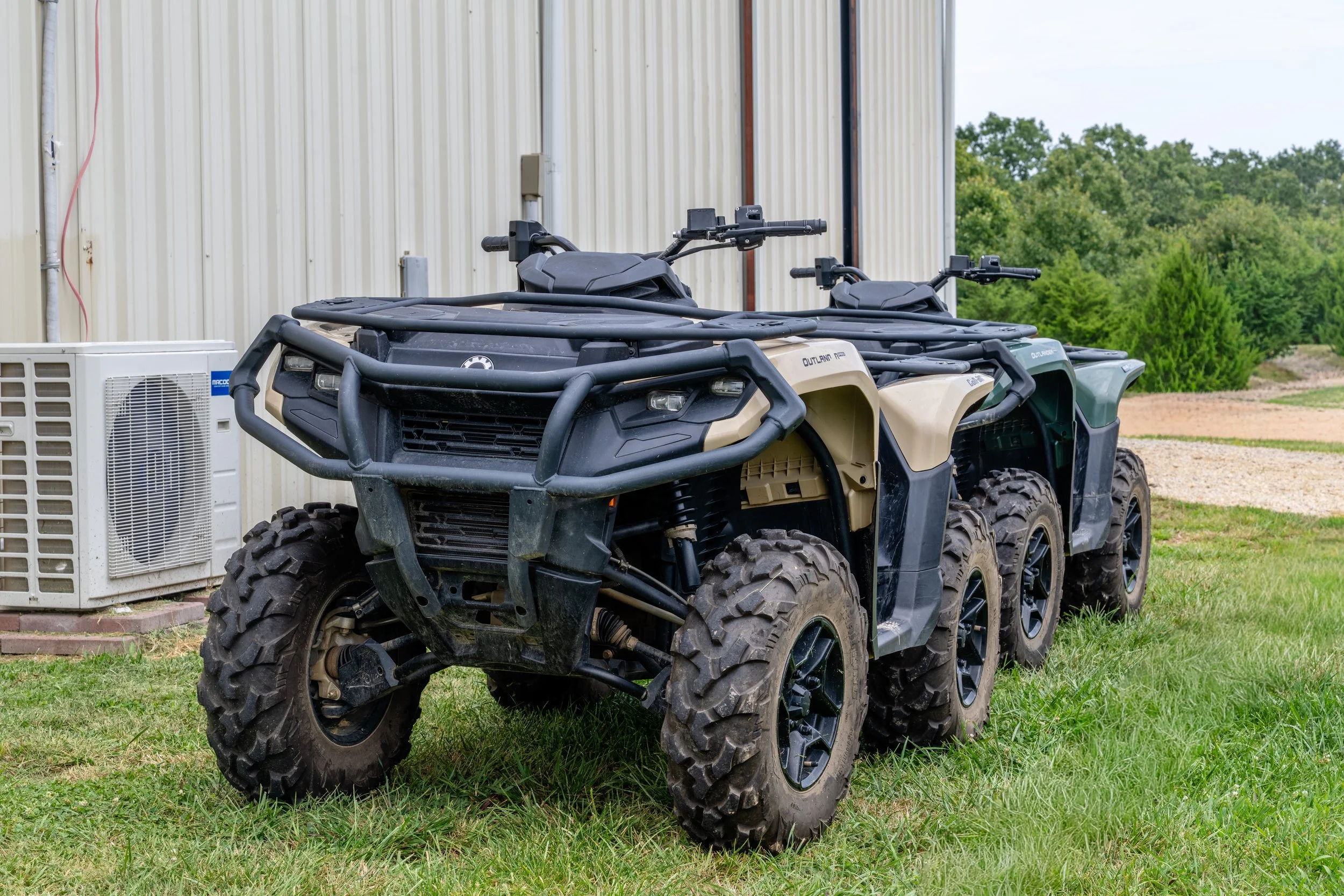 Two all-terrain vehicles parked on a grassy area next to a building, with an air conditioning unit nearby and trees in the background.