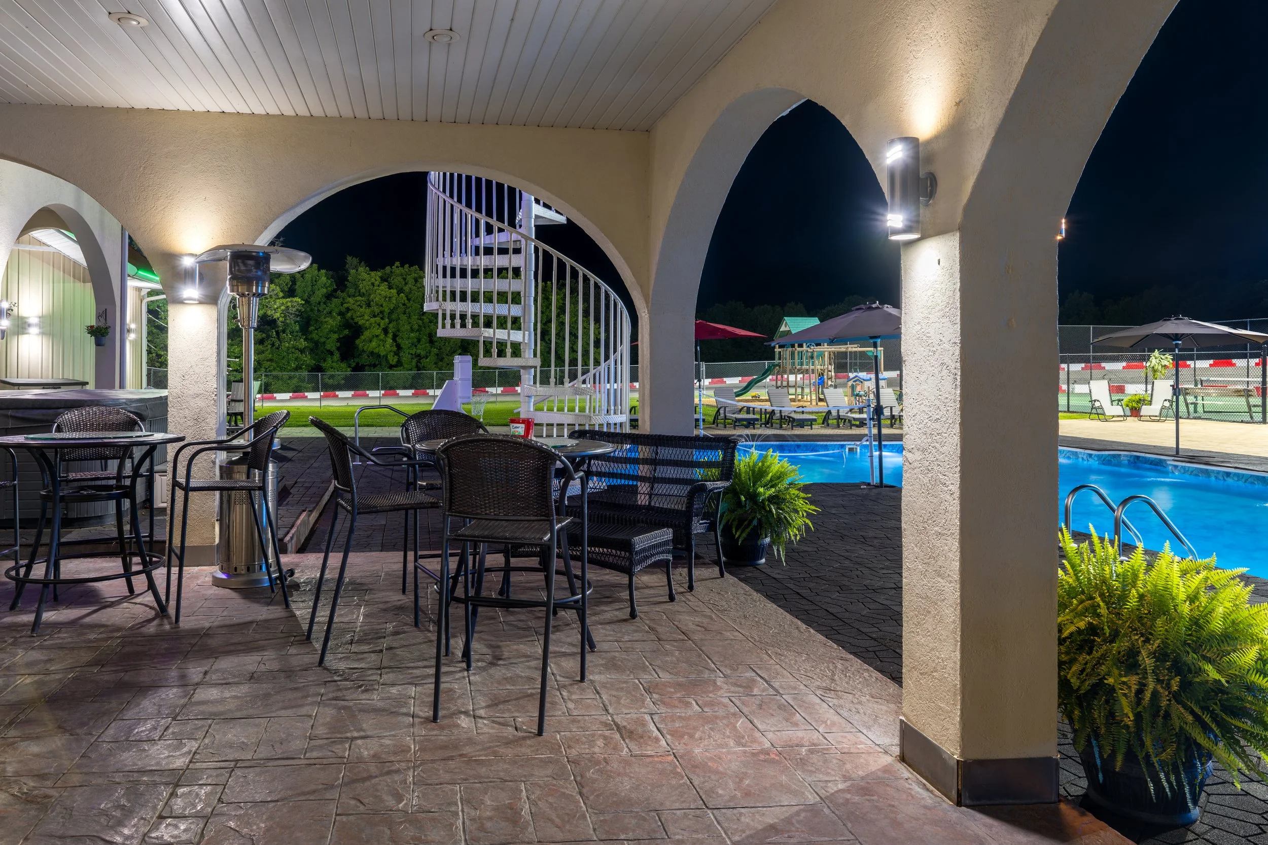 Night view of an outdoor pool area with lounge chairs, umbrellas, and a playground, seen from under a covered patio with arches.