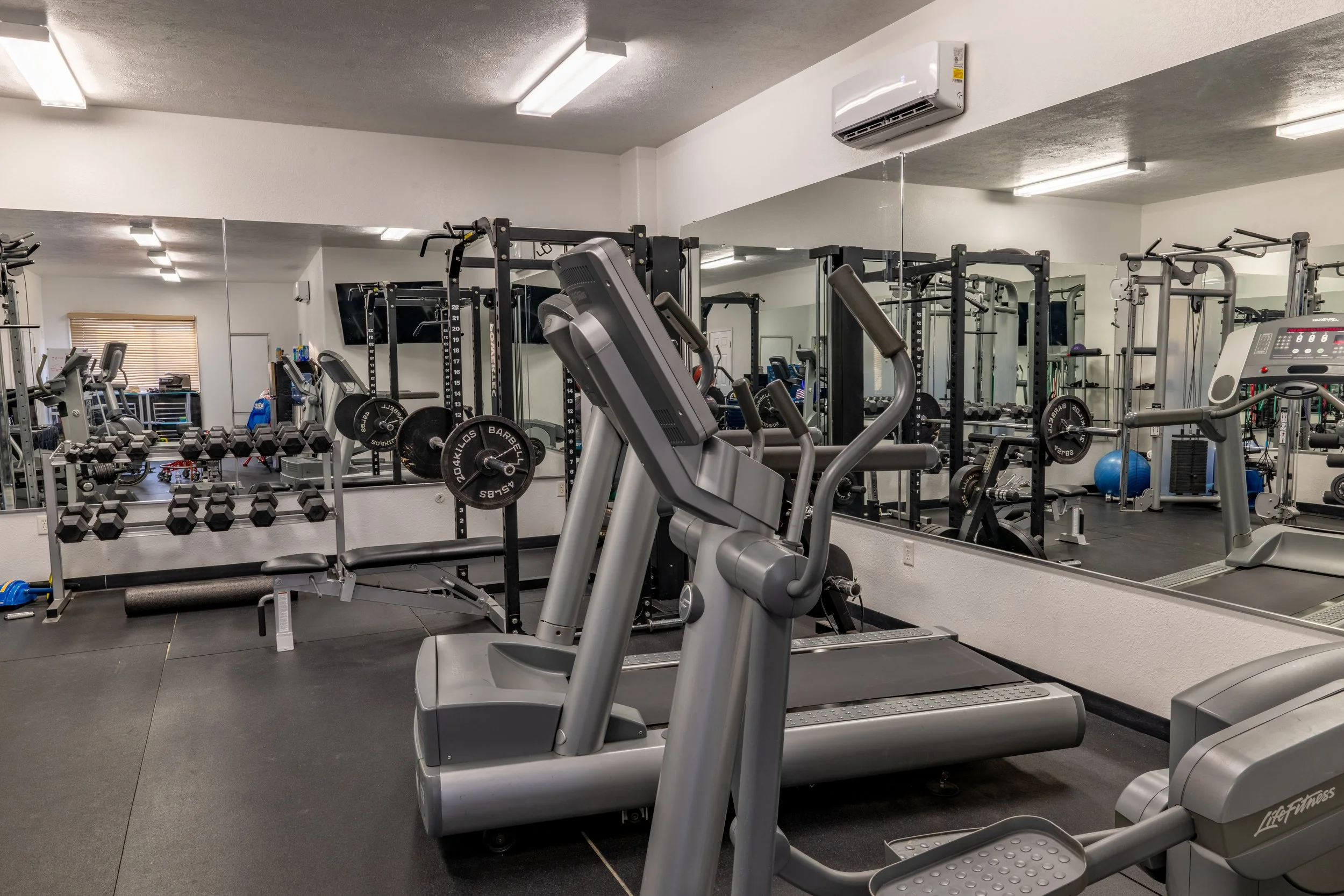 Interior of a gym with various exercise equipment including elliptical machines, dumbbells, and weightlifting stations, with a large mirror on one wall.