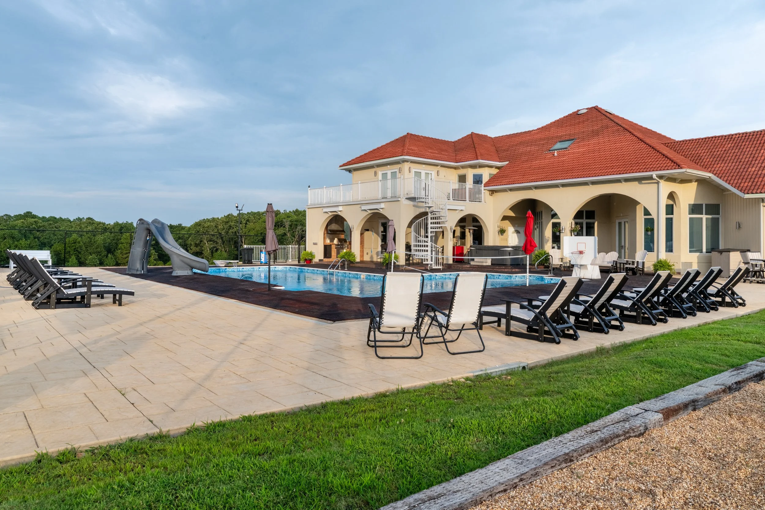 A backyard with a swimming pool surrounded by lounge chairs, umbrellas, and a house with a red tile roof, set against a cloudy sky.