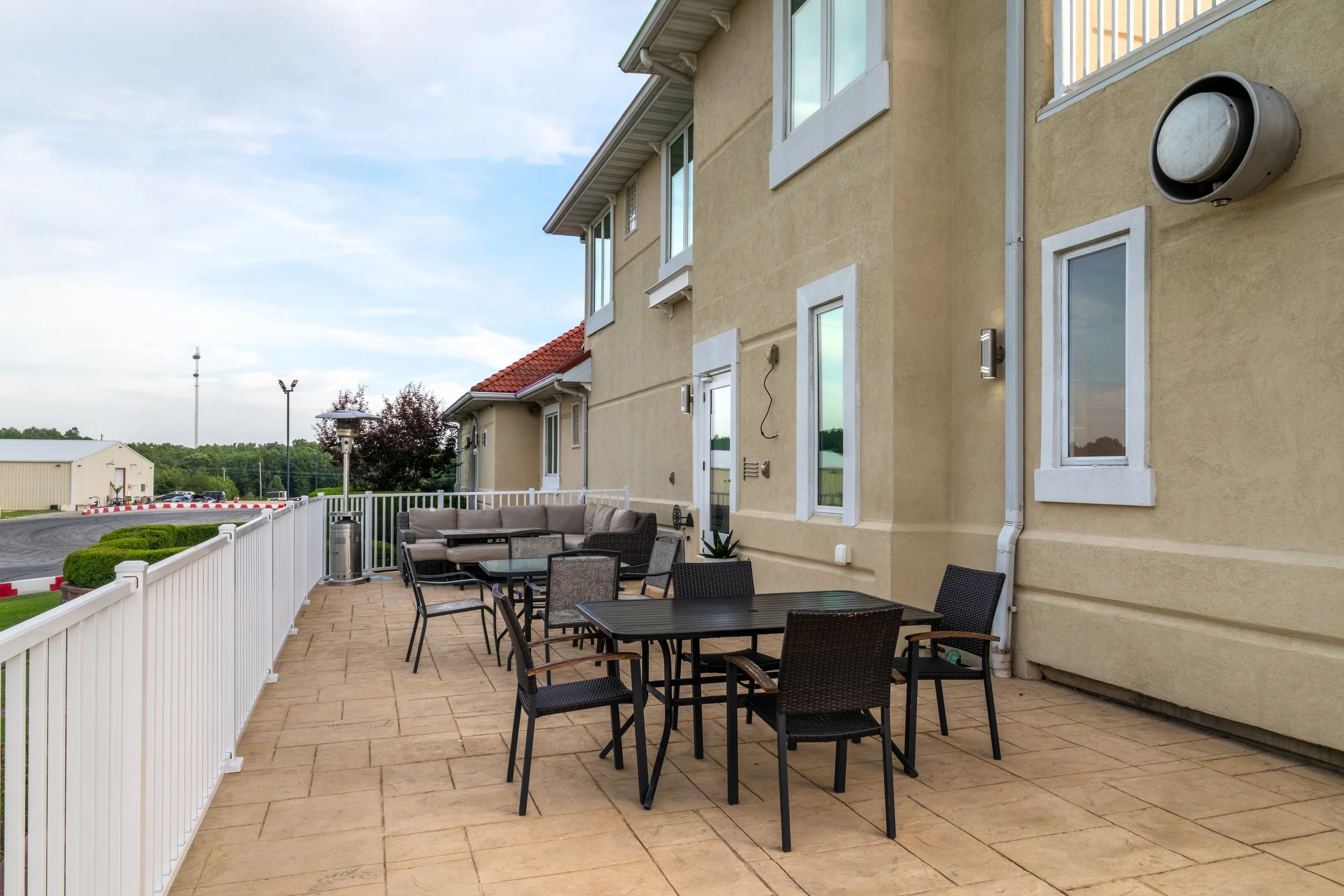 Outdoor patio area with black tables and chairs, beige stone flooring, beige stucco building with white window frames, wall-mounted lights, fencing, and some greenery in the background.
