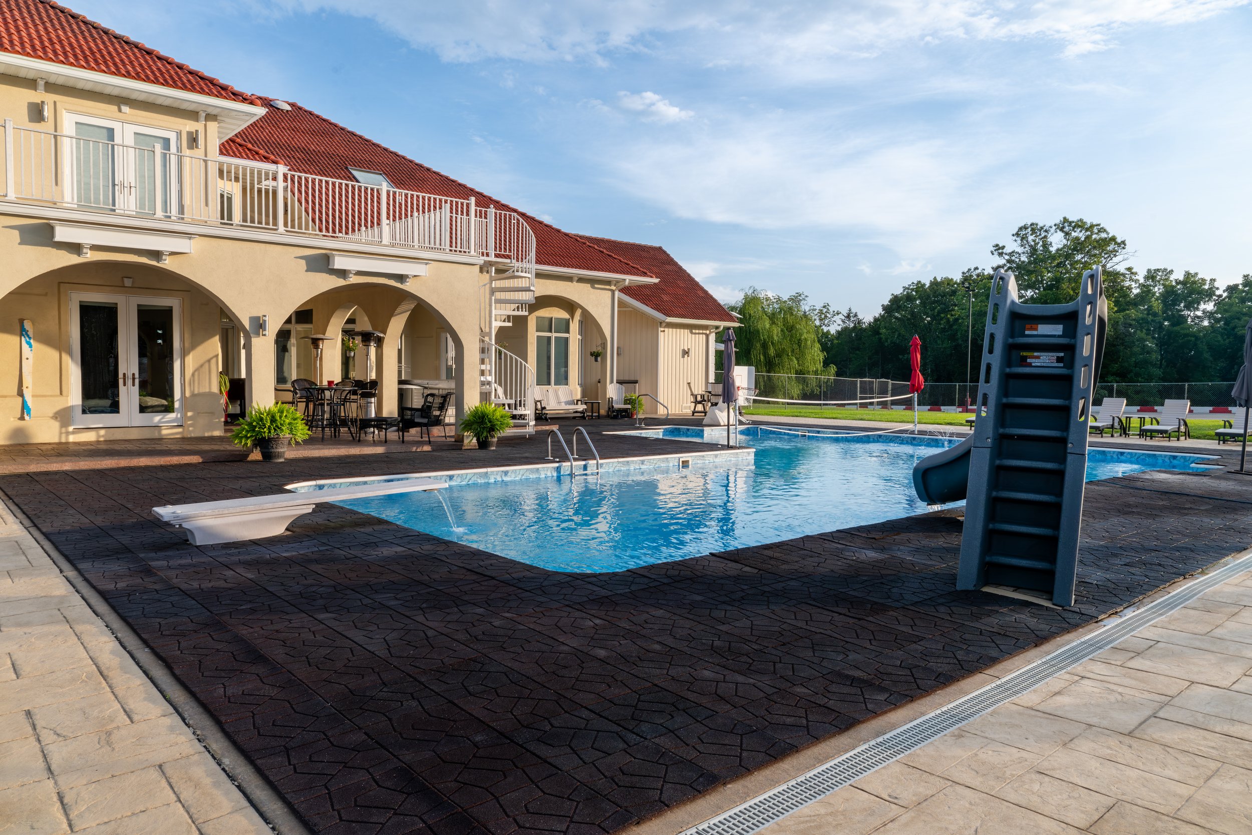 A swimming pool area next to a yellow building with a red-tiled roof, outdoor furniture, and umbrellas, with trees and a blue sky in the background.