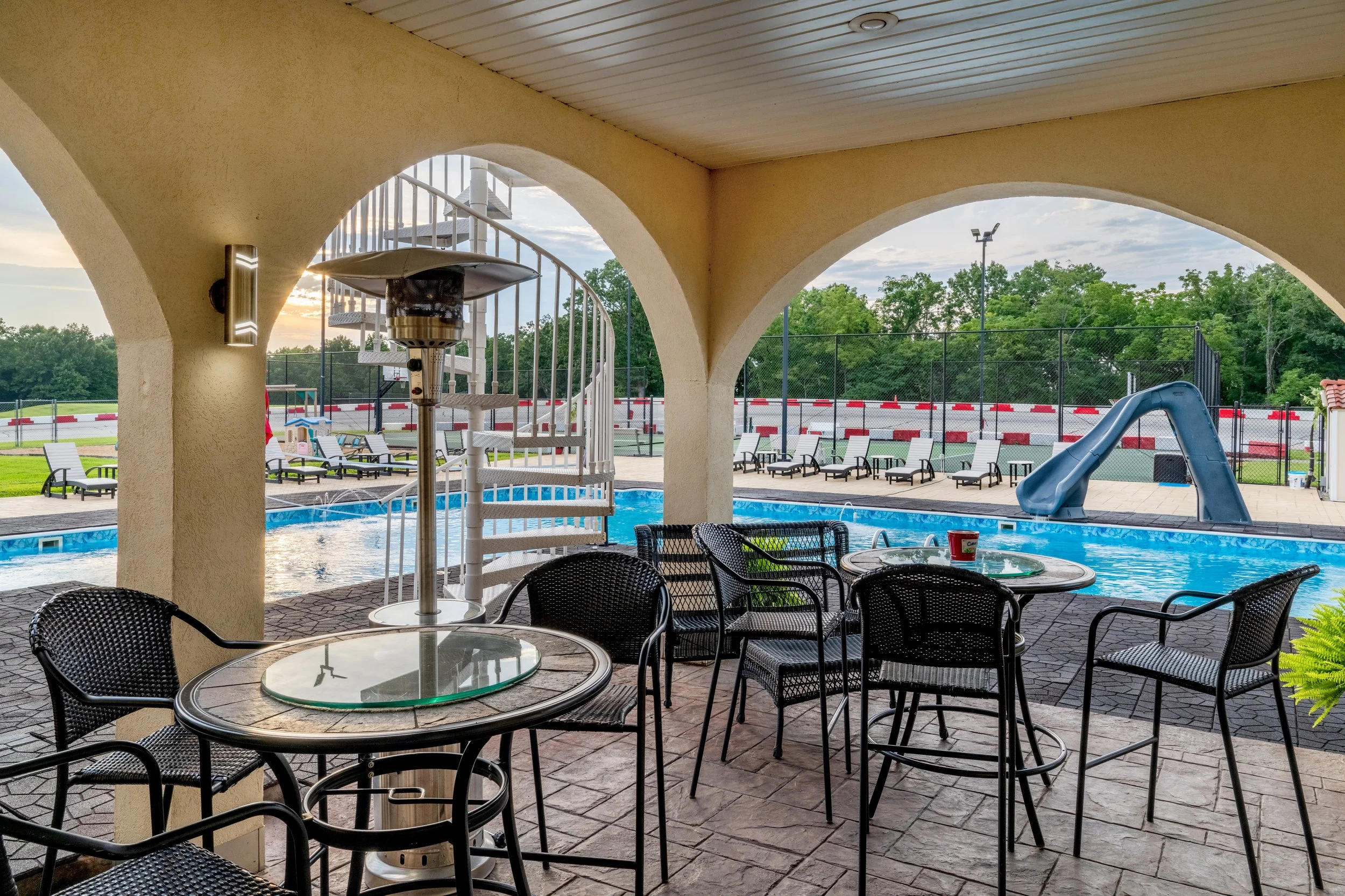 Poolside area with tables and chairs under shaded patio, swimming pool with slide in background, tennis court, trees, and a sunset sky.