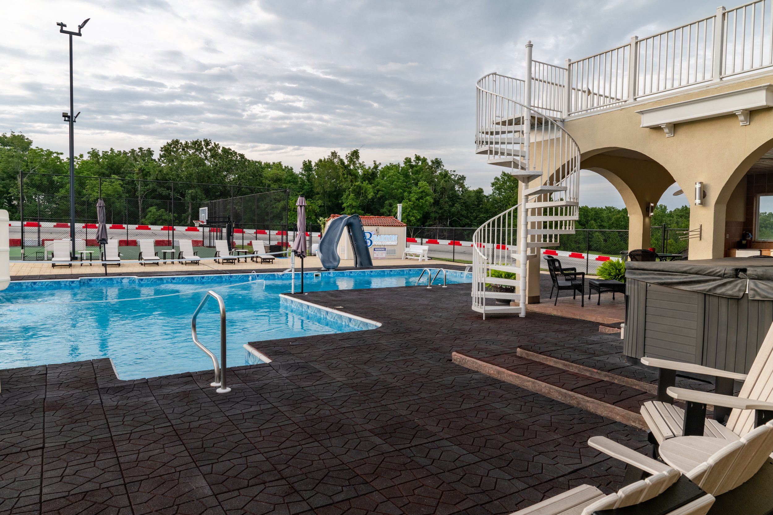 An outdoor swimming pool area with lounge chairs, umbrellas, a slide, and a fenced-in pool basketball hoop under a cloudy sky.
