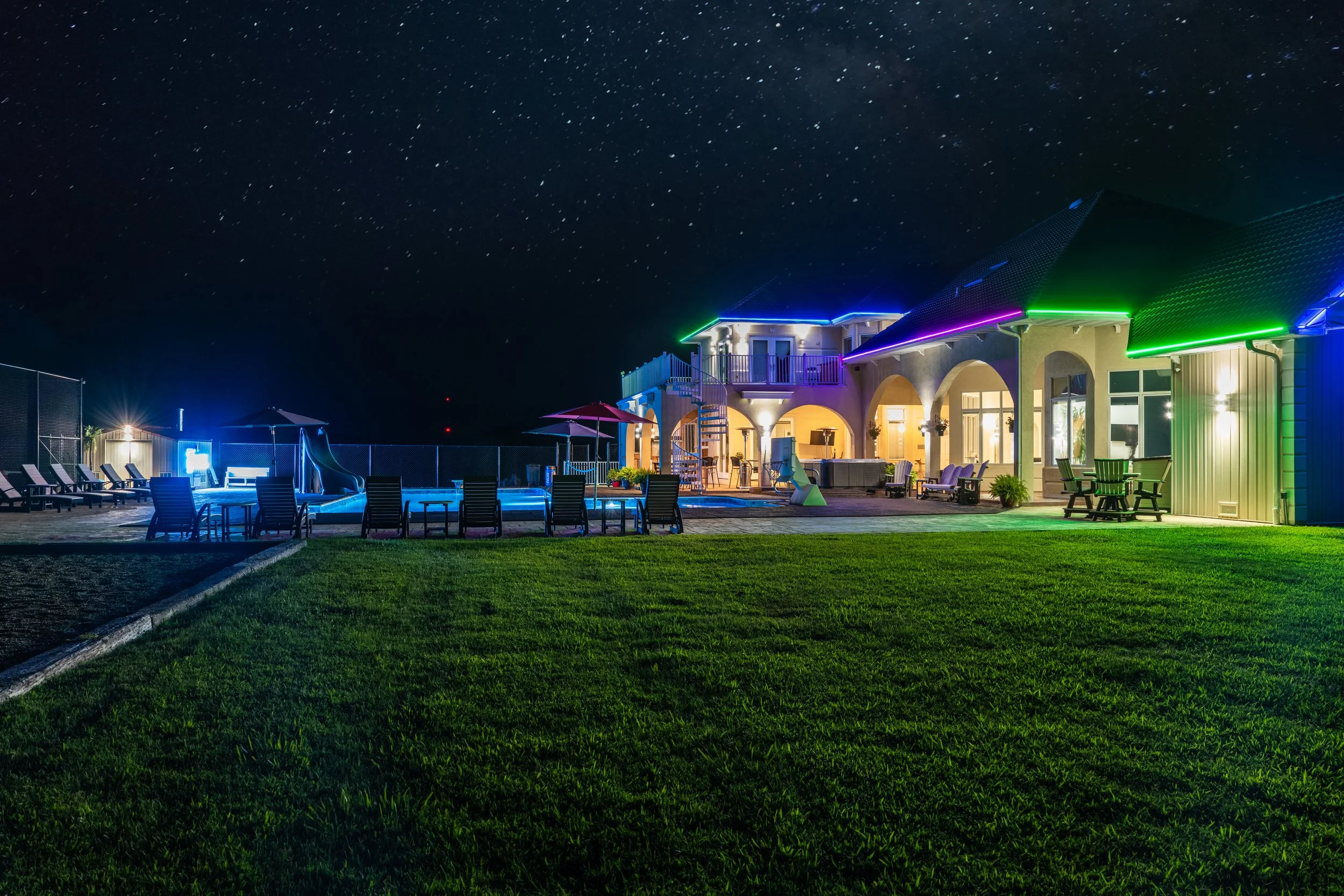 Night view of a house with colorful neon lighting, a swimming pool with lounge chairs, umbrellas, and a well-maintained lawn under a starry sky.