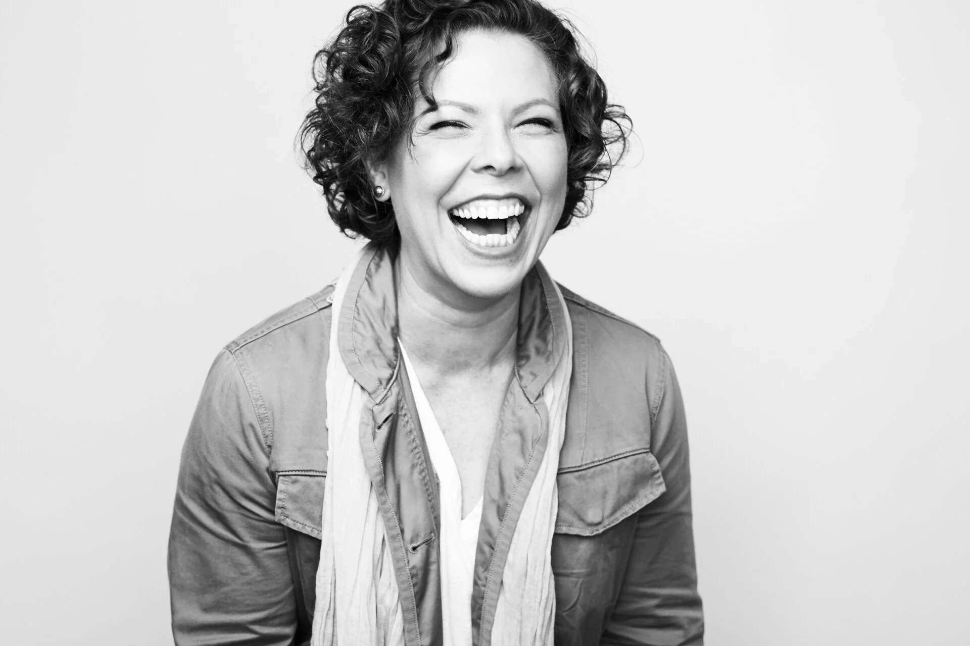 A woman with short, curly hair laughing with her eyes nearly closed, wearing a denim jacket and earrings, against a plain light background.