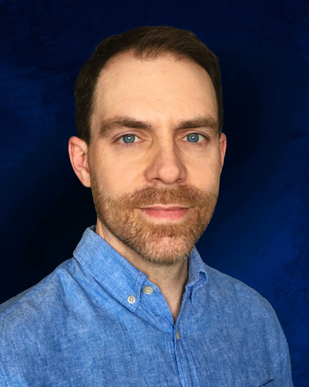 A portrait of a man with blue eyes, brown hair, and a beard, wearing a light blue button-up shirt against a dark blue background.