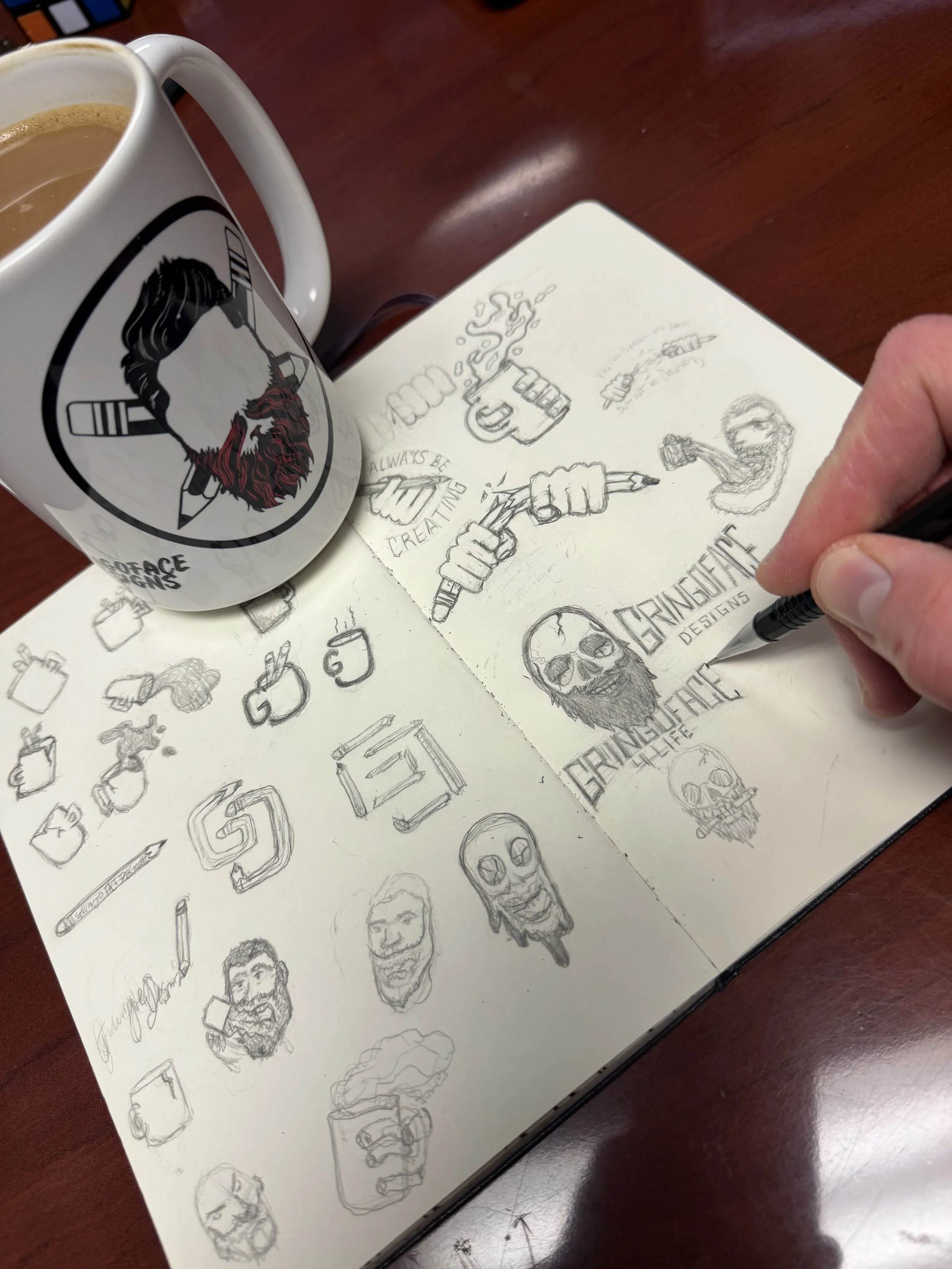 Sketched illustrations and logos on a notebook, a coffee mug with a logo of a bearded man, coffee, and a hand in the process of drawing.