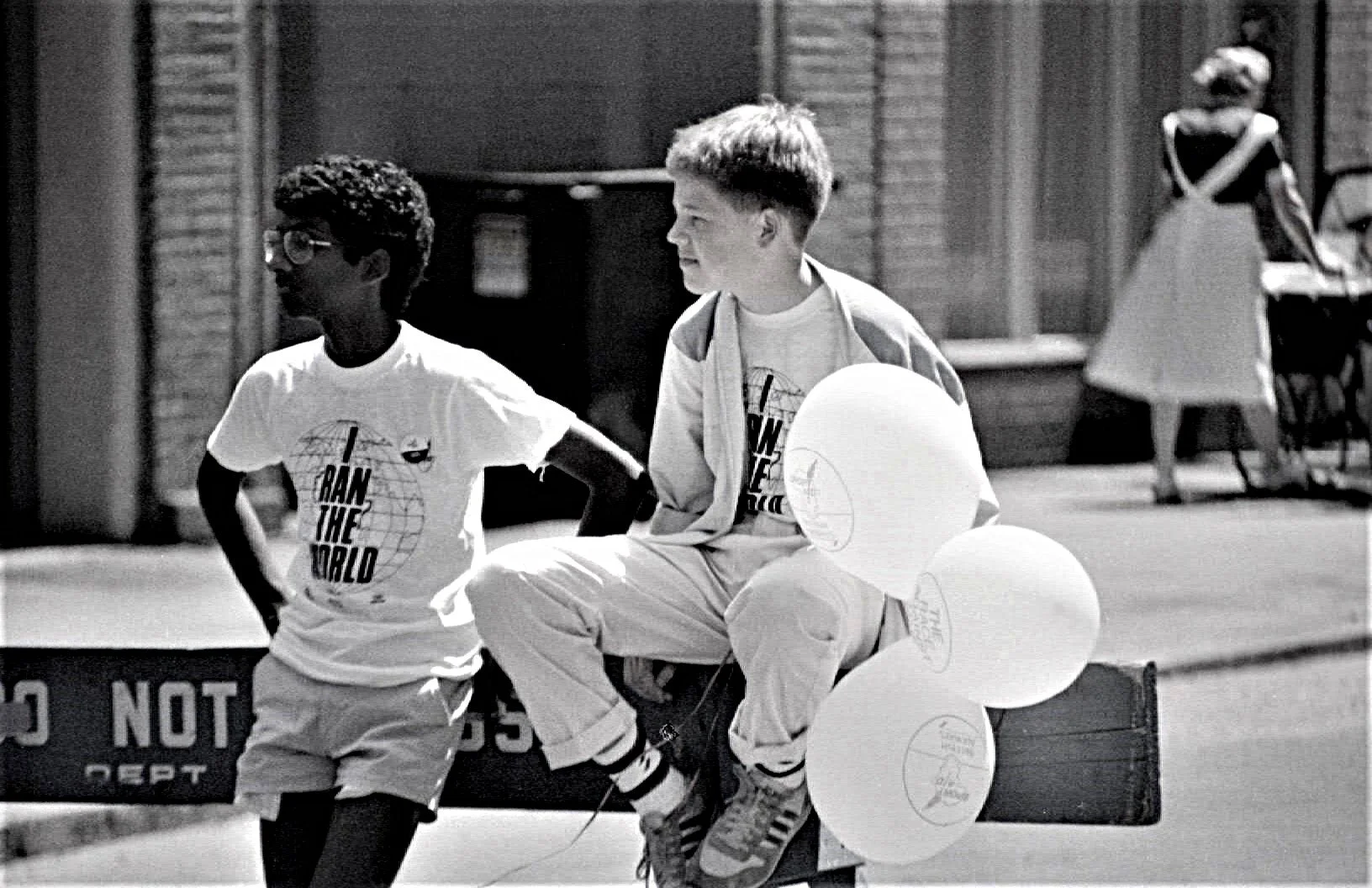 Two New York young boys sitting and standing on a bench outside, one wearing glasses and a T-shirt with the text "Rain the World" and the other wearing a jacket, holding balloons, with a woman in a dress and a man with a bicycle in the background.