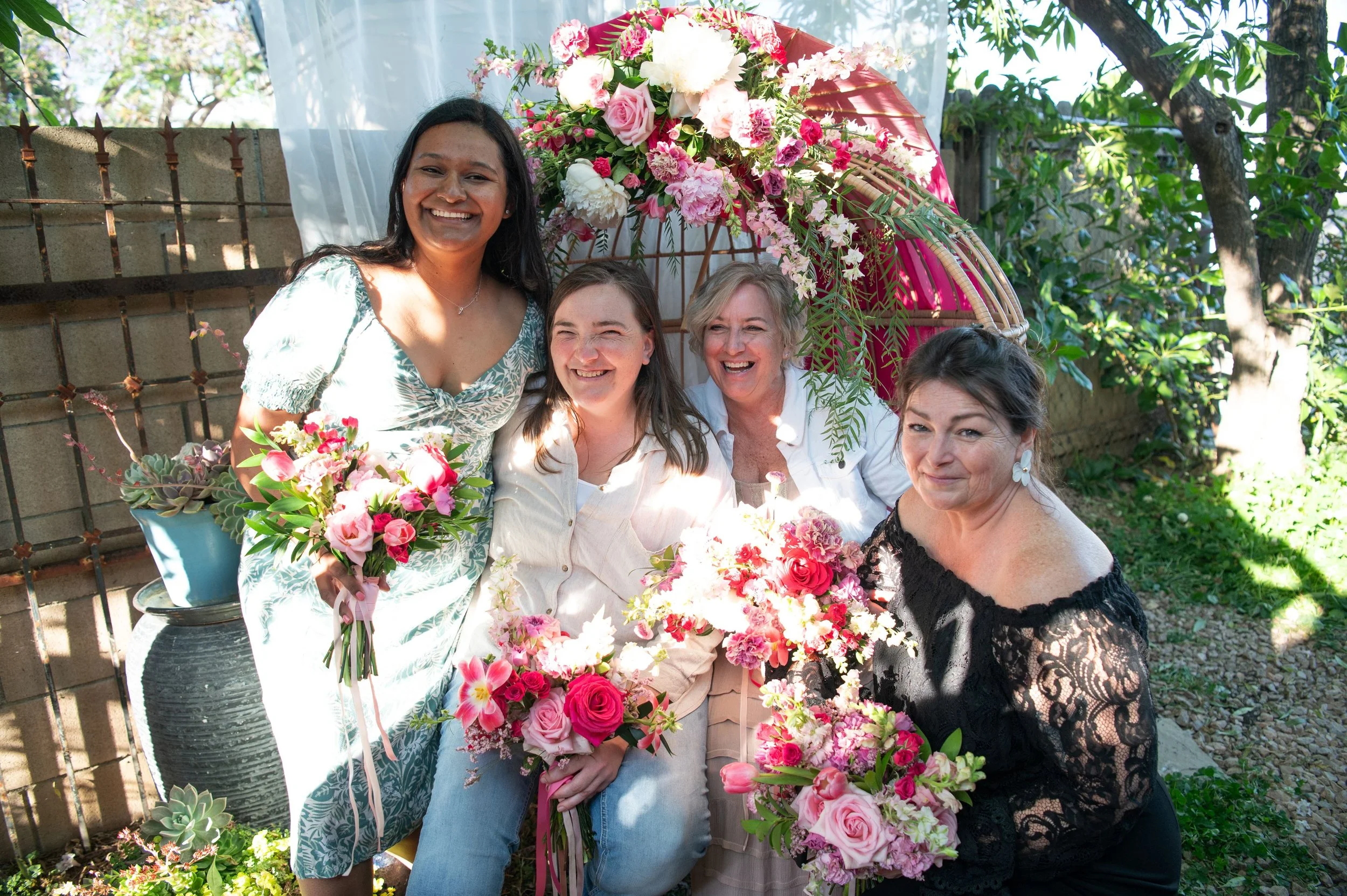 A group of four women smiling and posing outdoors at a flower-themed gathering, surrounded by pink and white flowers on a large floral arrangement and in pots, with trees and greenery in the background.