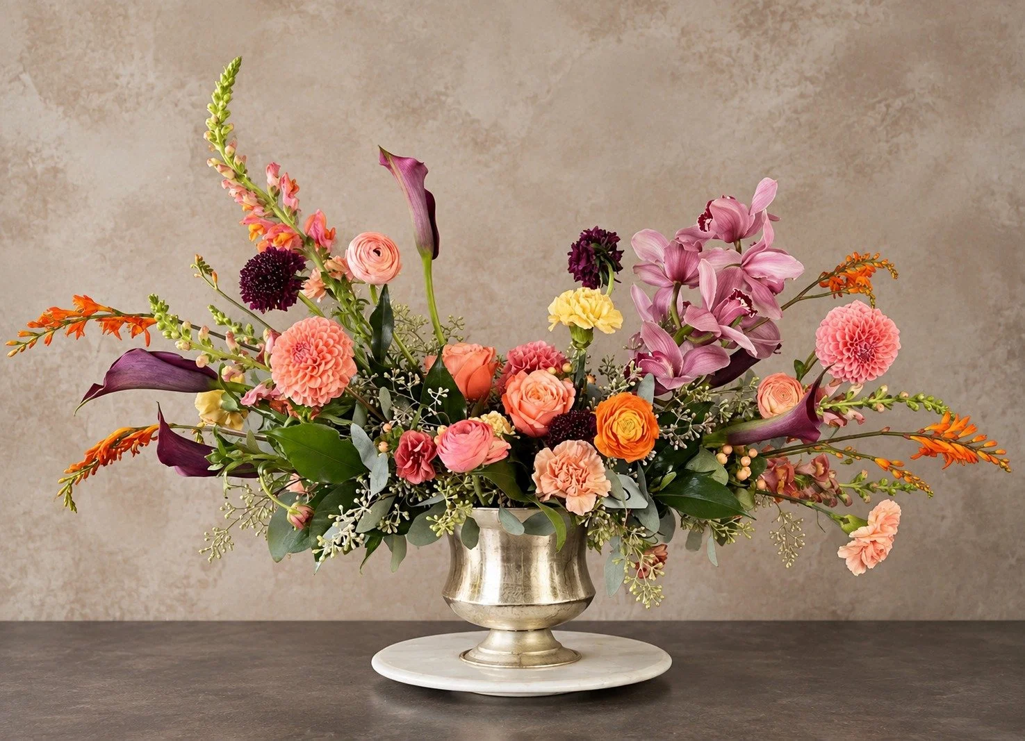 Large autumn floral arrangement with orchids, calla lilies, peach and orange roses, plum flowers and sweeping seasonal botanicals in a silver vase.