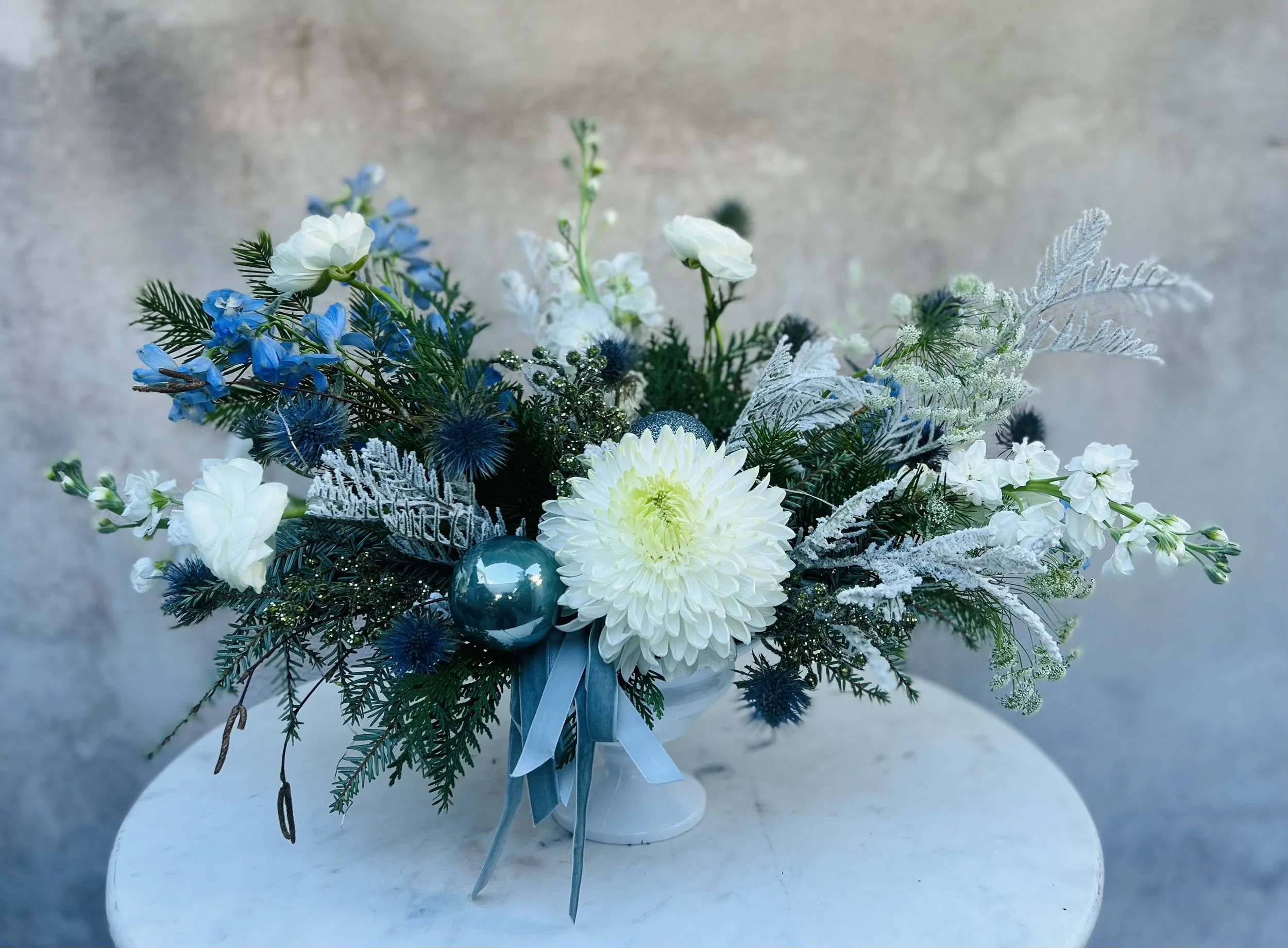 Medium-sized winter flower arrangement with white chrysanthemums, blue delphinium, blue thistle, ornaments, and lush winter greens in a stylish white vase on a white table with a gray background.