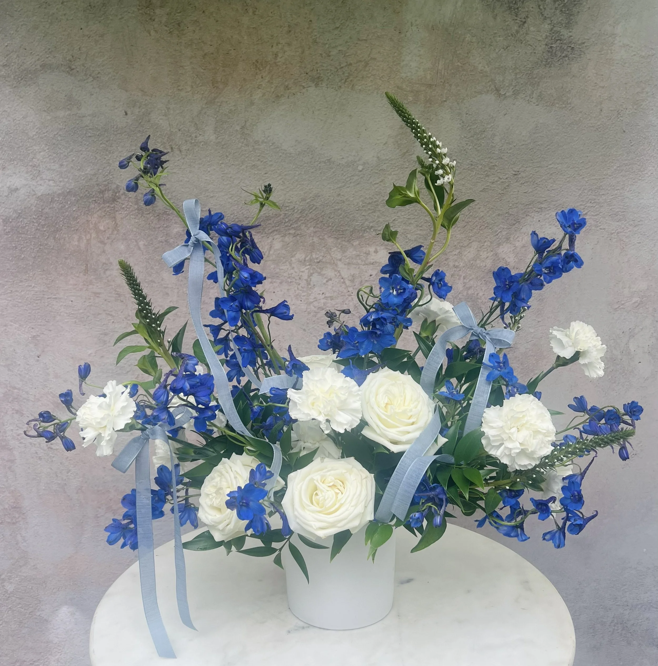 Soft white and vibrant blue flower arrangement in a glass vase featuring seasonal blooms and greenery designed for a new baby boy