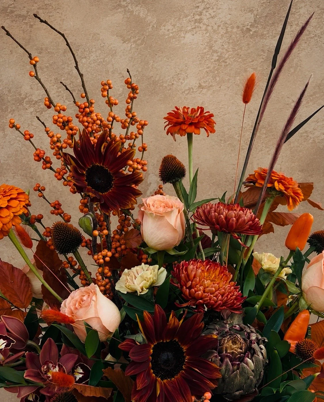 Close-up of burgundy sunflowers, peach roses, autumn chrysanthemums, orange berries and artichoke in a richly textured fall arrangement.