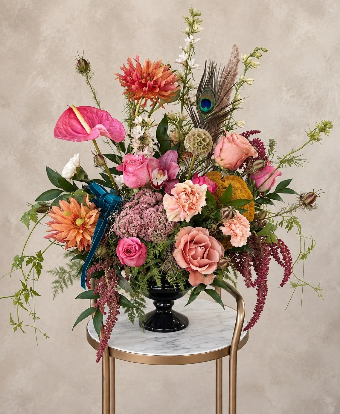 Artistic autumn floral arrangement with pink anthurium, roses, peach blooms, burgundy amaranthus, greenery and a peacock feather in a black compote vase.