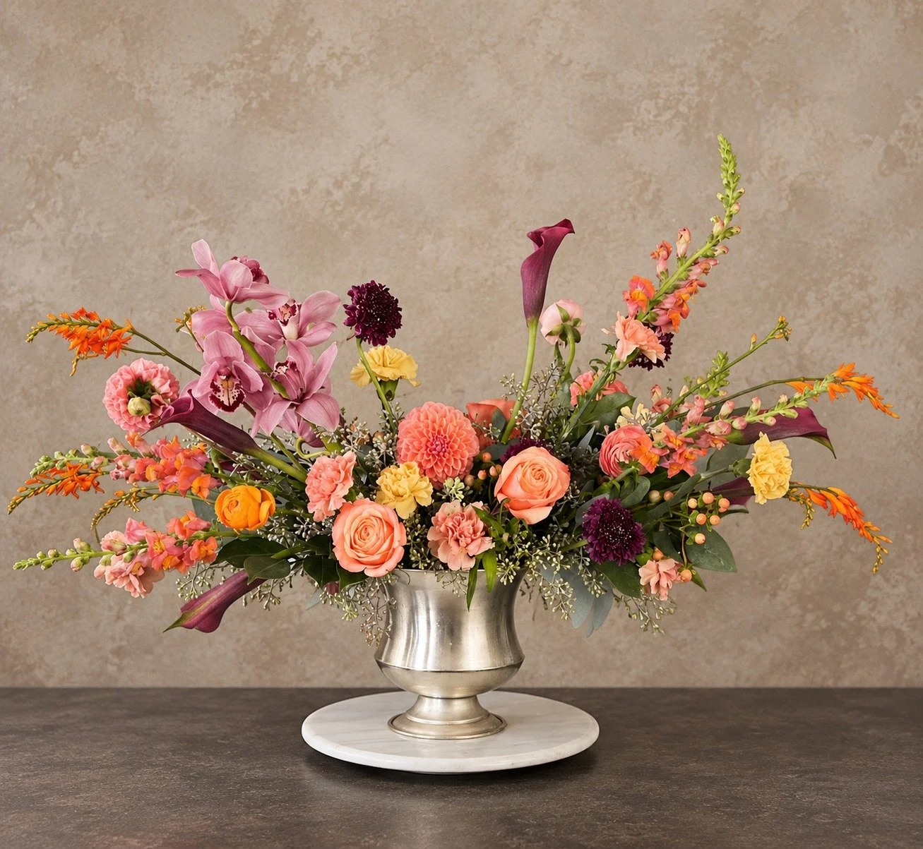 Luxury autumn statement arrangement with pink orchids, plum calla lilies, peach roses, dahlias and seasonal flowers in a silver pedestal vase.