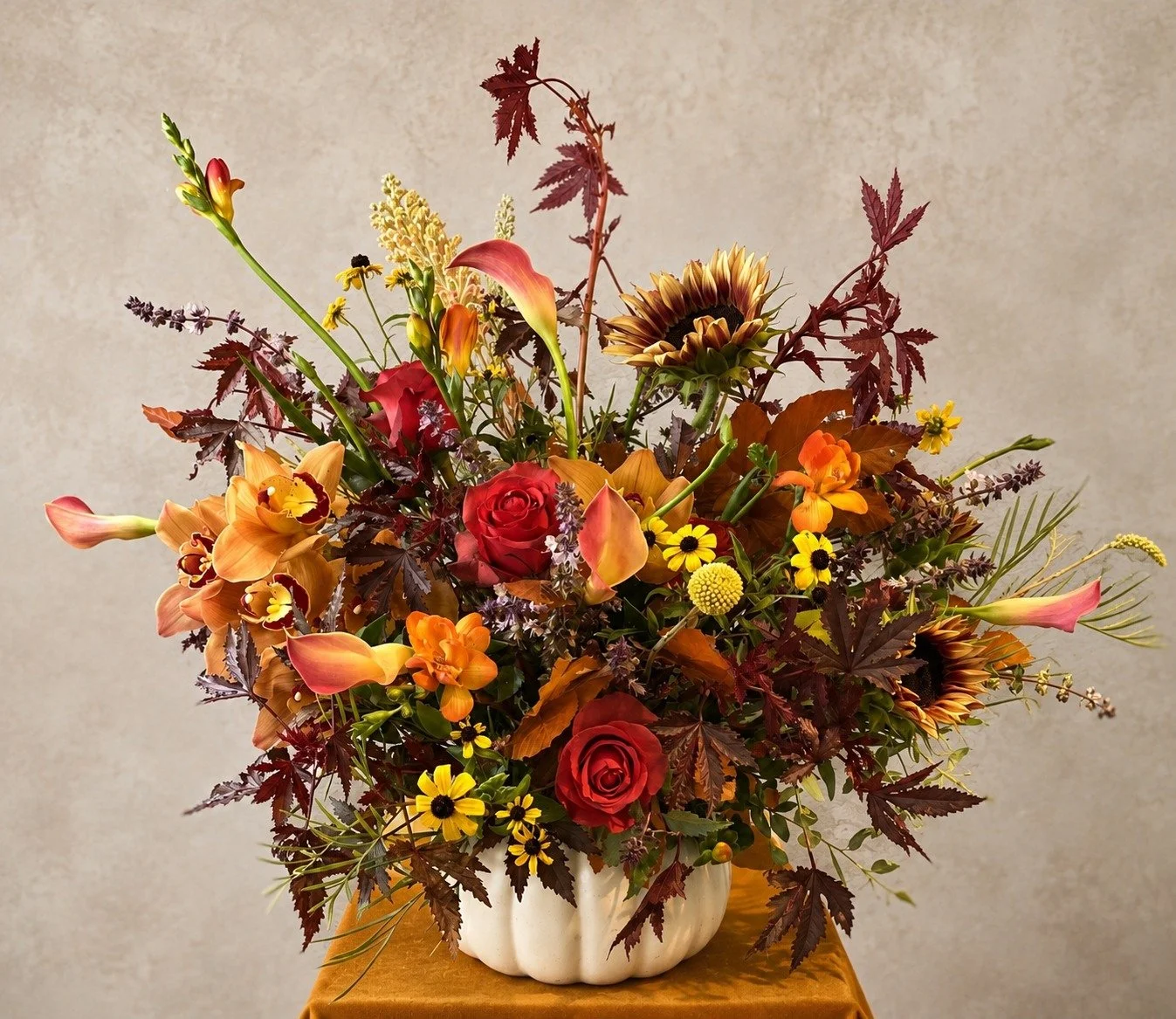 Autumn floral arrangement with orange orchids, red roses, calla lilies, sunflowers and burgundy foliage in a white pumpkin vase.