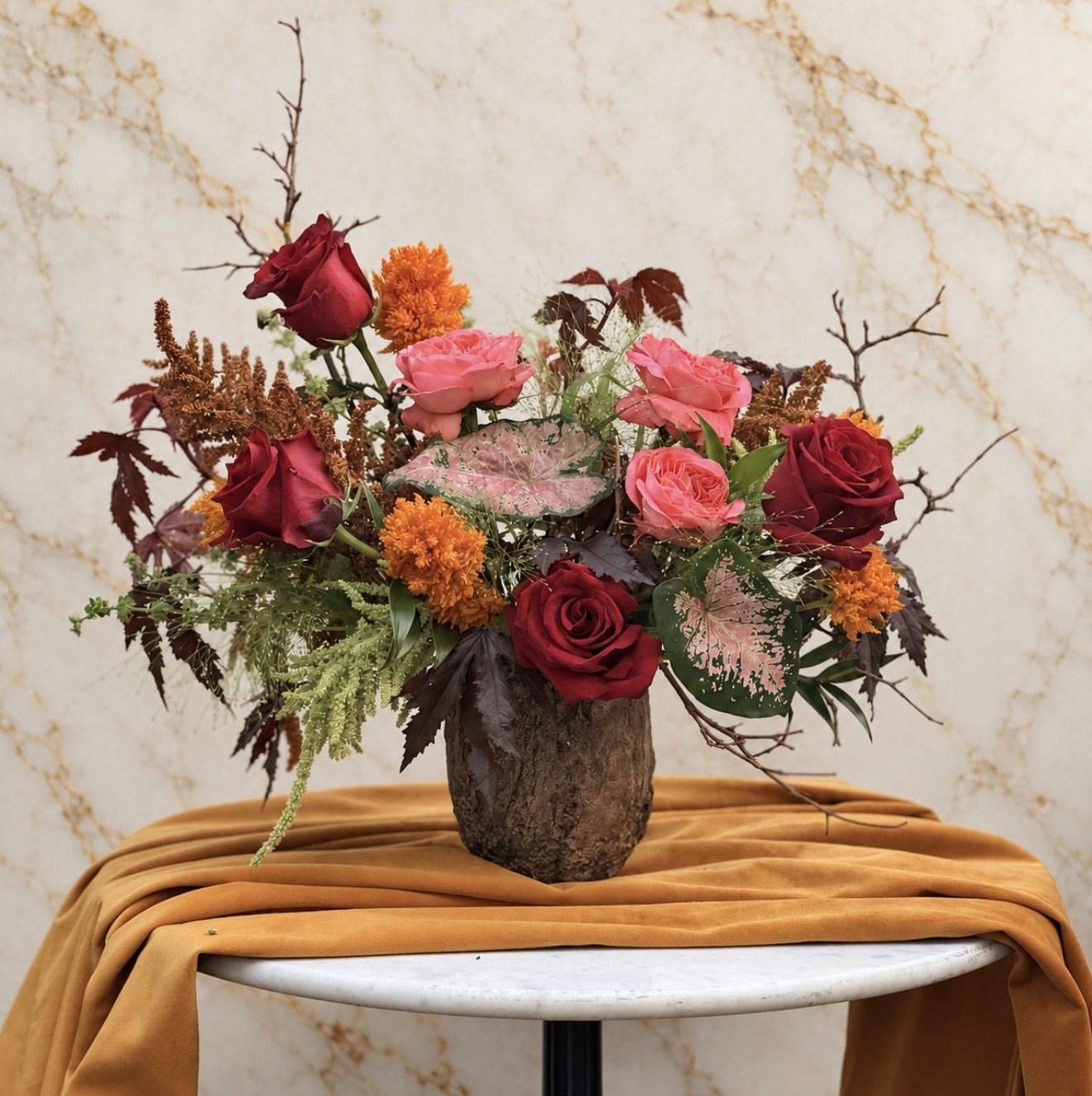 Moody autumn floral arrangement with burgundy and pink roses, orange blooms and seasonal foliage in a dark textured vase.