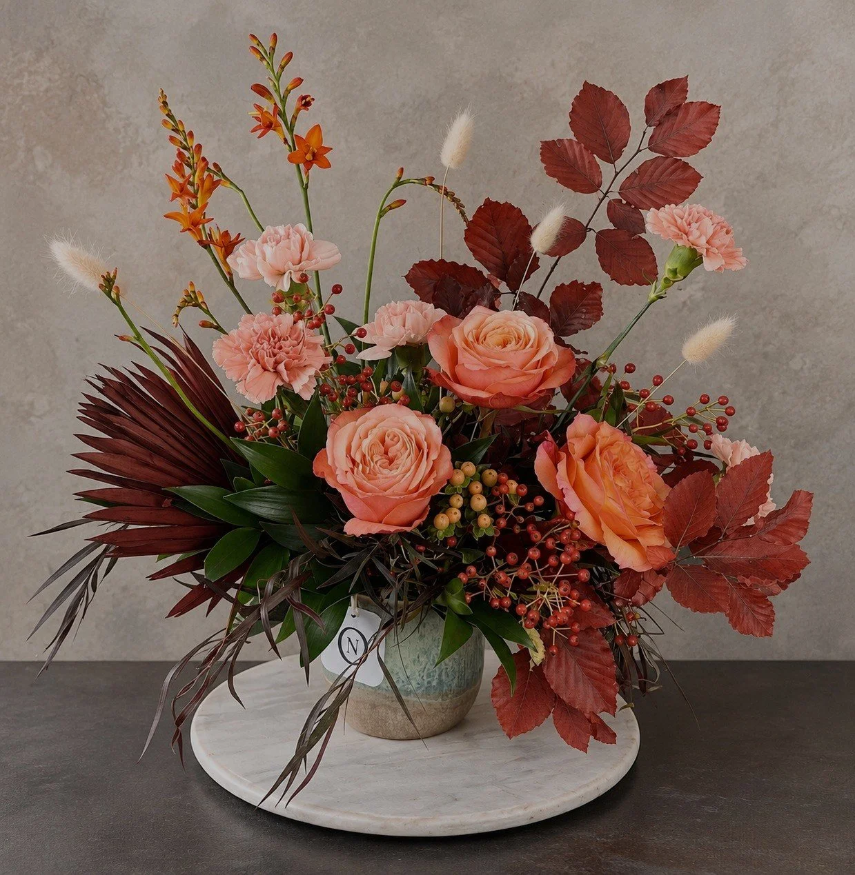 Refined autumn floral arrangement with peach roses, blush carnations, burgundy foliage, berries and seasonal grasses in a ceramic vase.