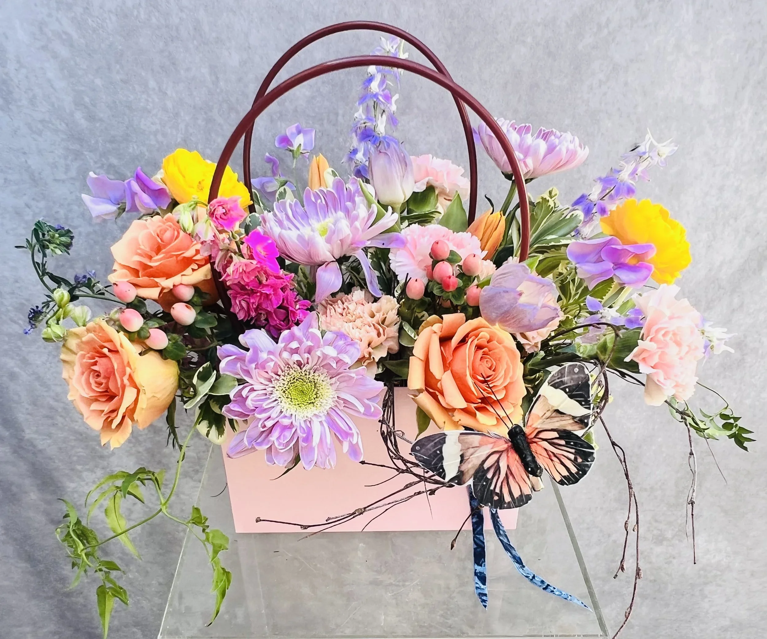 Pastel floral arrangement with seasonal flowers and greenery in a pink keepsake kraft tote bag.