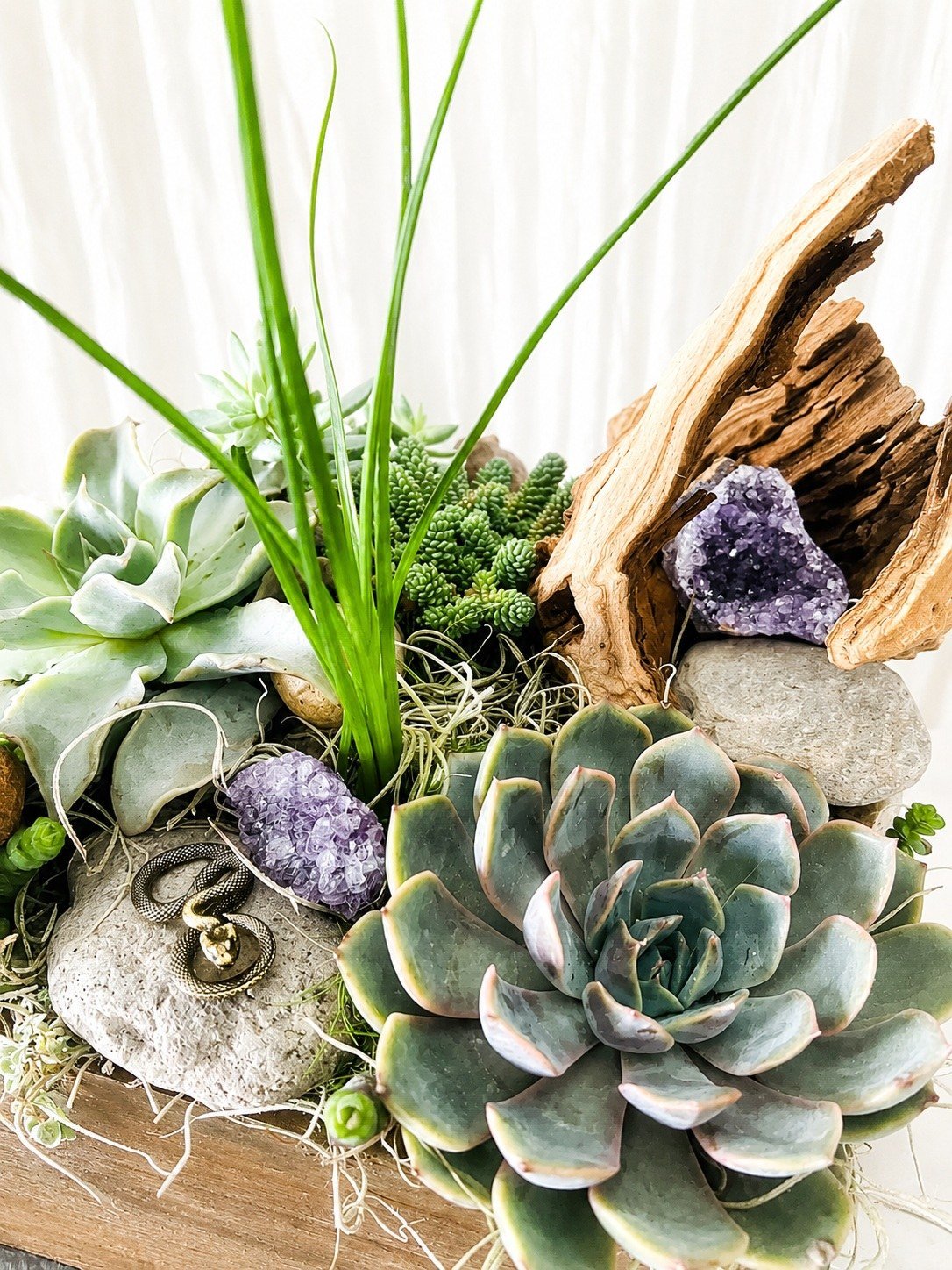 Close-up of a handcrafted living succulent garden featuring sculptural succulents, natural driftwood, lush greenery, and amethyst crystals arranged in a handmade square wood planter.
