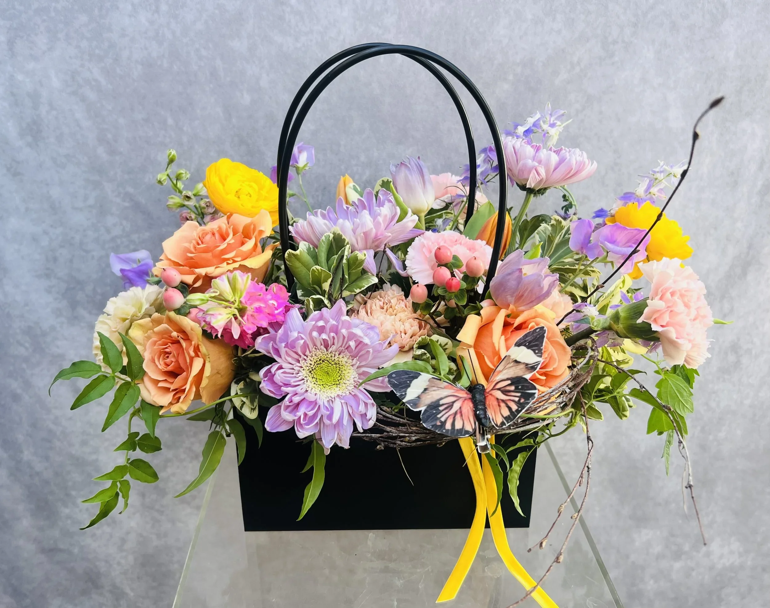 Pastel floral arrangement with seasonal flowers and greenery in a black keepsake kraft tote bag.