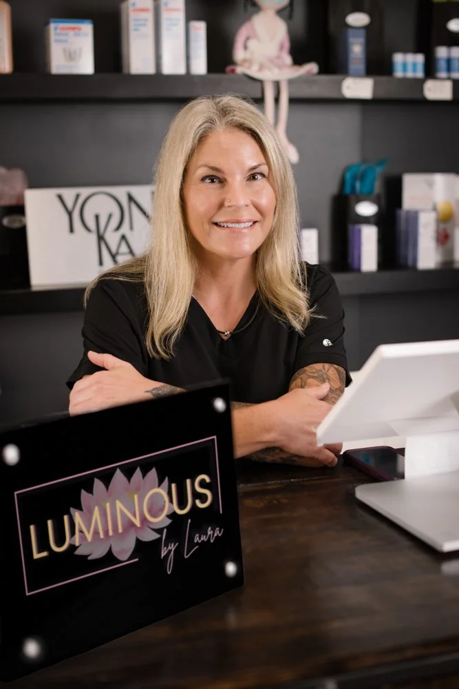 Smiling woman with blonde hair sitting at a desk in a retail or office environment, holding a clipboard, with a sign on the desk that says 'LUMINOUS by Laura'.